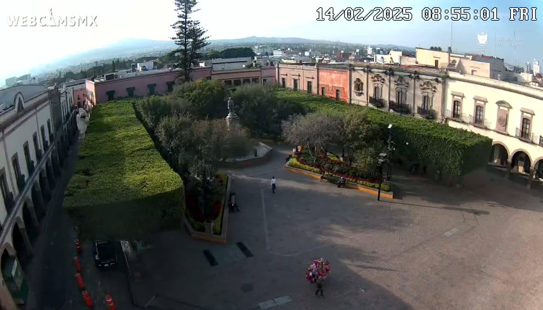 A high-angle view of a plaza, on a sunny day, surrounded by colonial-style buildings and neatly trimmed hedges, with a few people visible.
