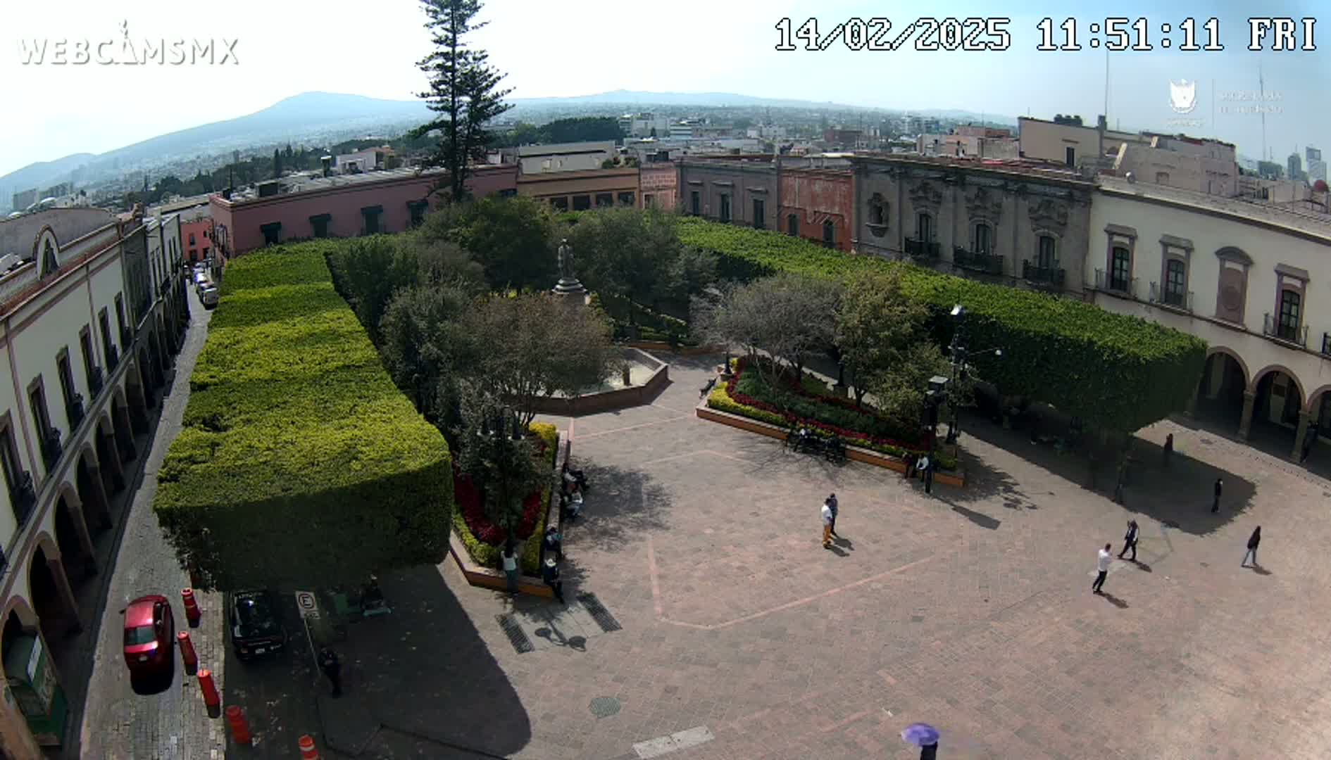 A town square with a central fountain, surrounded by neatly trimmed hedges, buildings, and a few people, under a sunny sky.