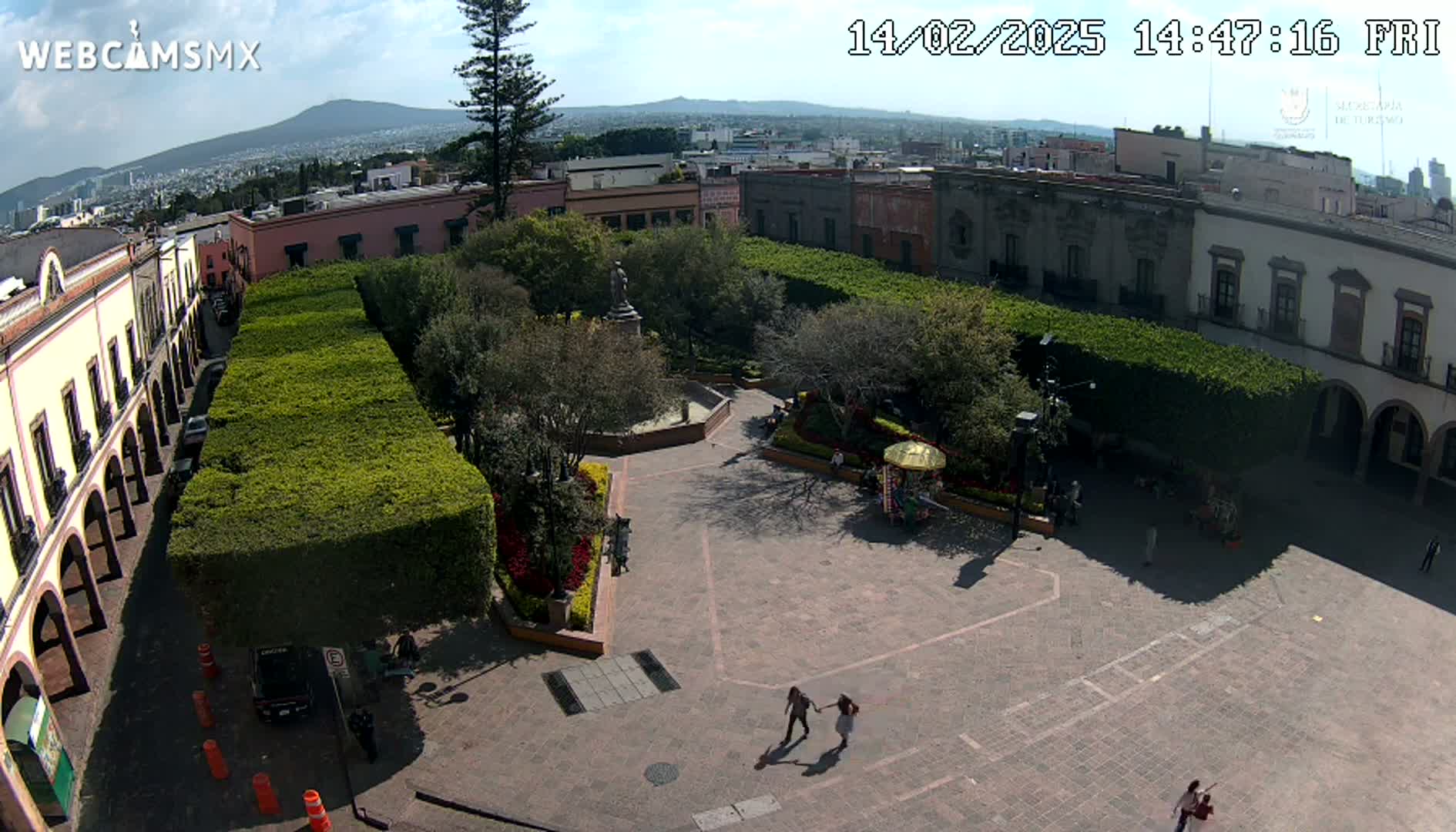 A sunny aerial view shows a town square with buildings surrounding a paved plaza, green hedges, and a few people walking.