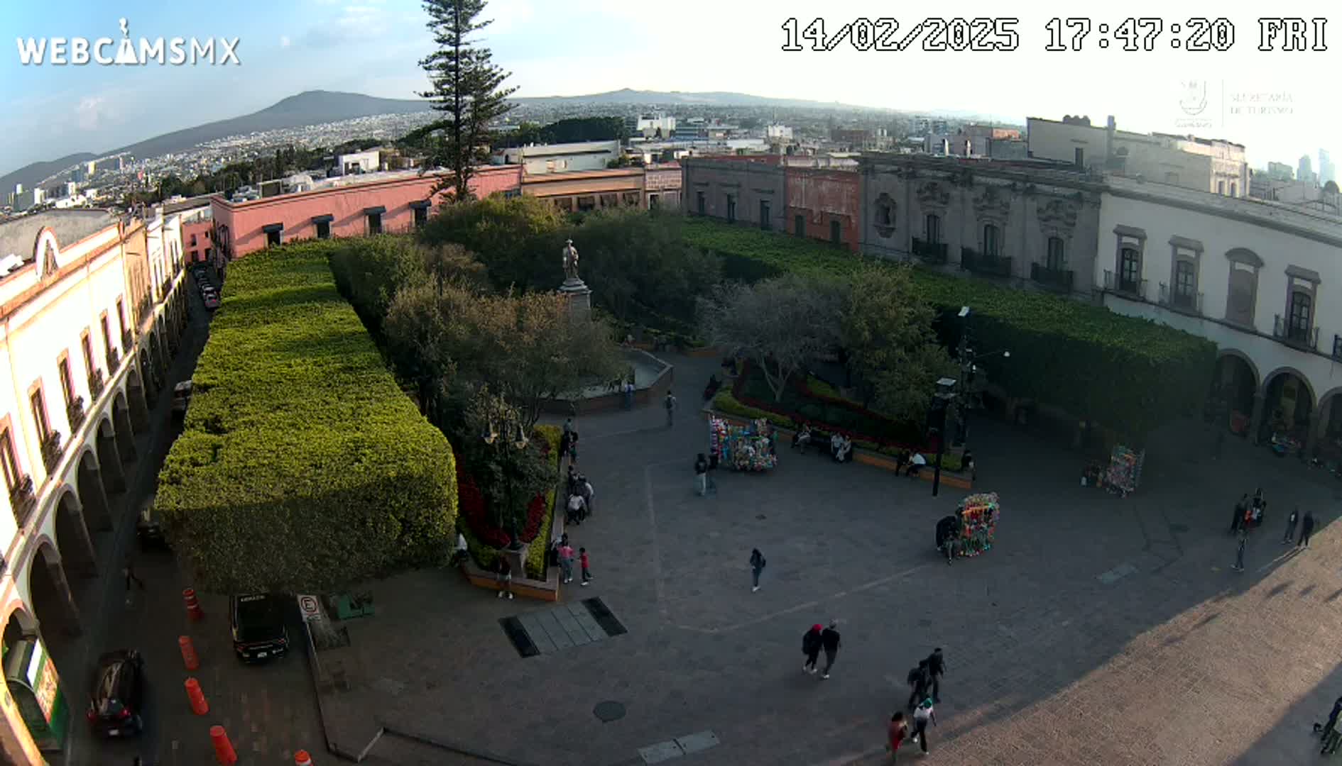 An aerial view of a town square, under partly sunny skies, shows people walking around a plaza with buildings lining its edges and lush green hedges in the center.