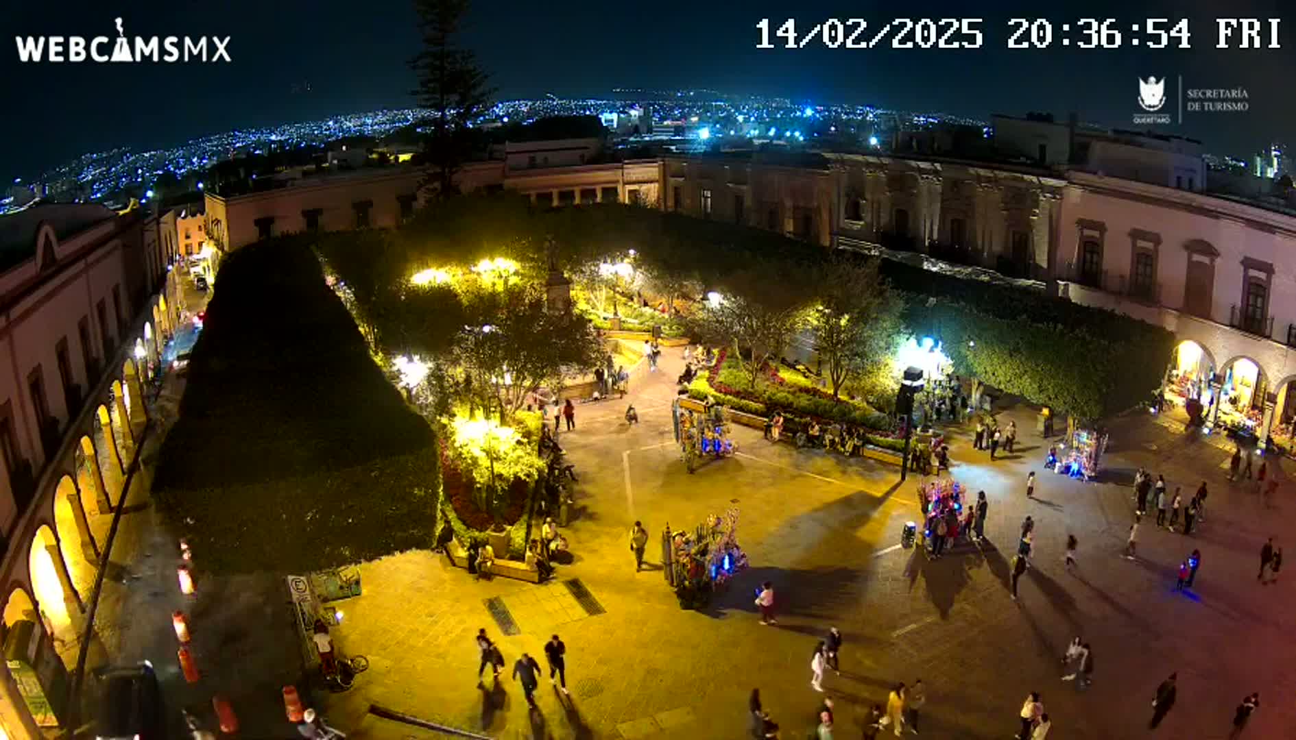 A nighttime aerial view of a brightly lit town square with people walking around, surrounded by buildings and lush green landscaping under a clear sky.