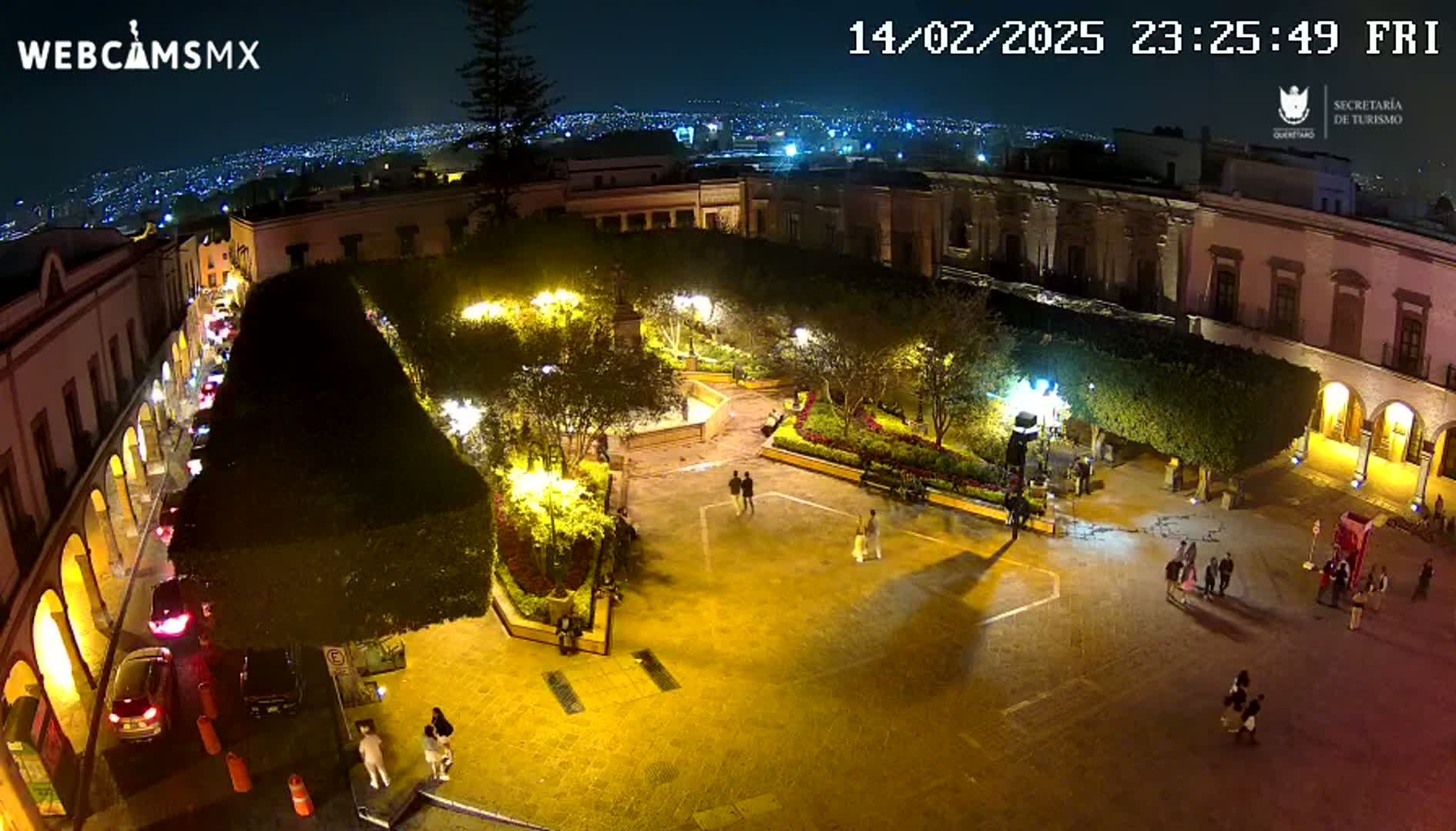 A nighttime aerial view shows a brightly lit plaza surrounded by colonial-style buildings, with several people walking in the square and cars parked along a street.