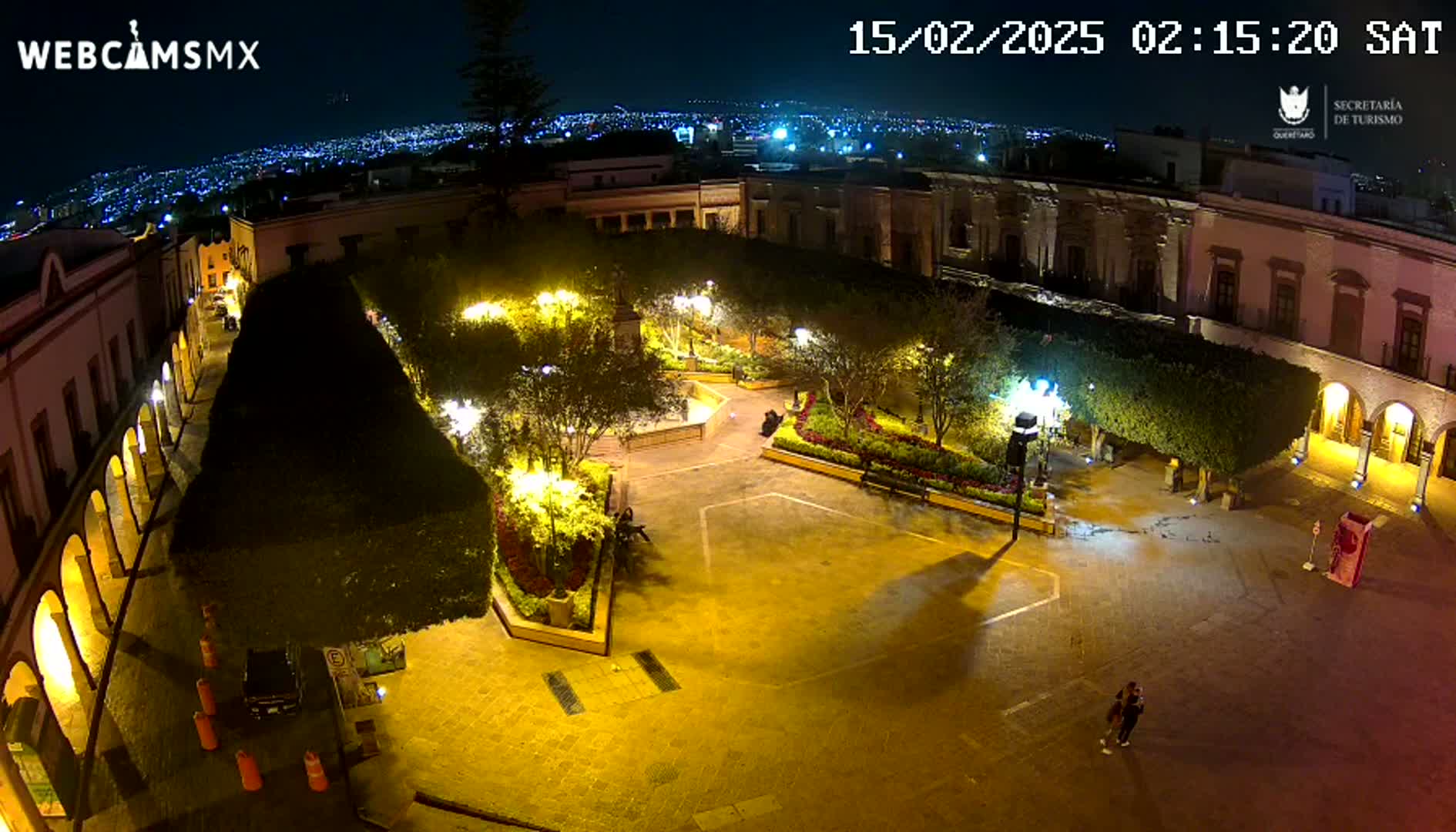 An illuminated plaza at night, surrounded by buildings, features landscaping, and a couple standing near the edge.