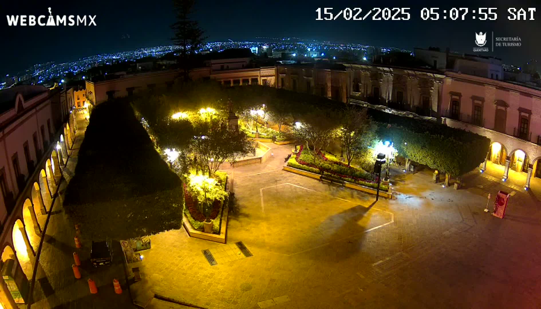 An illuminated plaza at night, surrounded by buildings, overlooks a city skyline under a clear sky.
