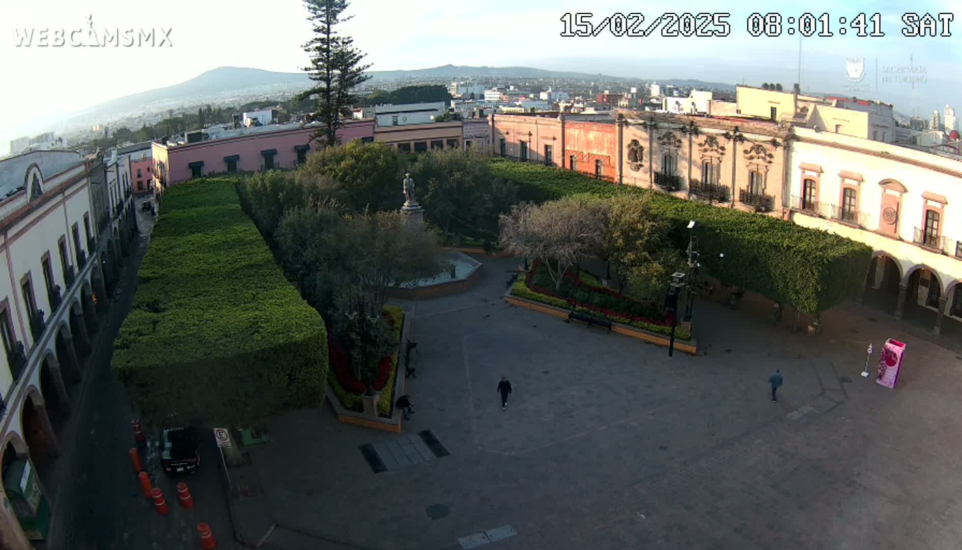 A mostly empty plaza with trimmed hedges, a fountain, and colonial-style buildings surrounds it under a clear sky.