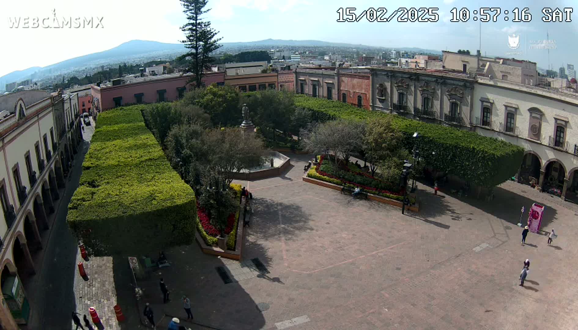 An elevated view of a town square with a central fountain, surrounded by neatly trimmed hedges and colonial-style buildings, under a sunny sky.