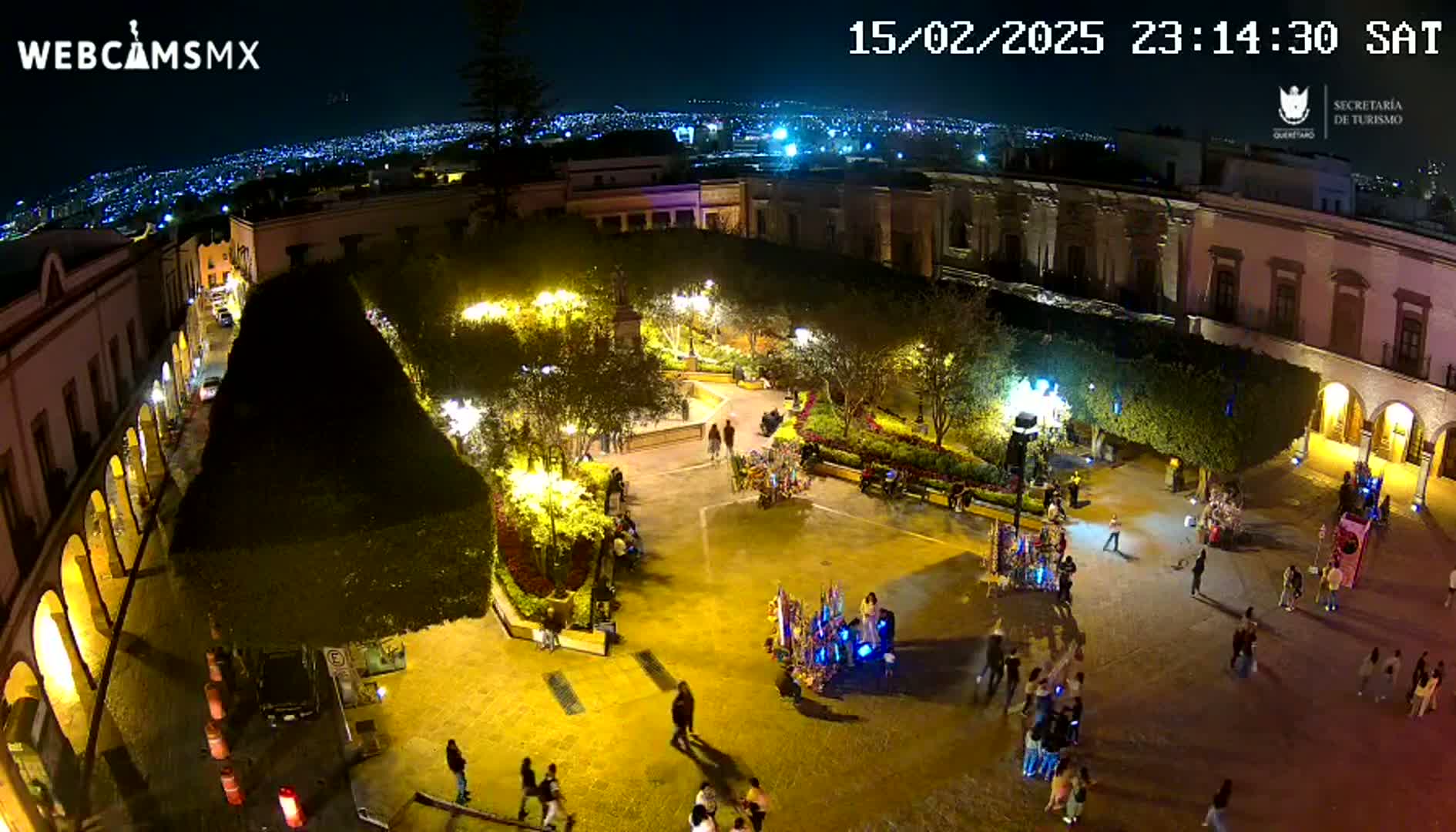 An illuminated town square at night, with people strolling around and small vendor stalls set up, is overlooked by buildings under a clear sky.