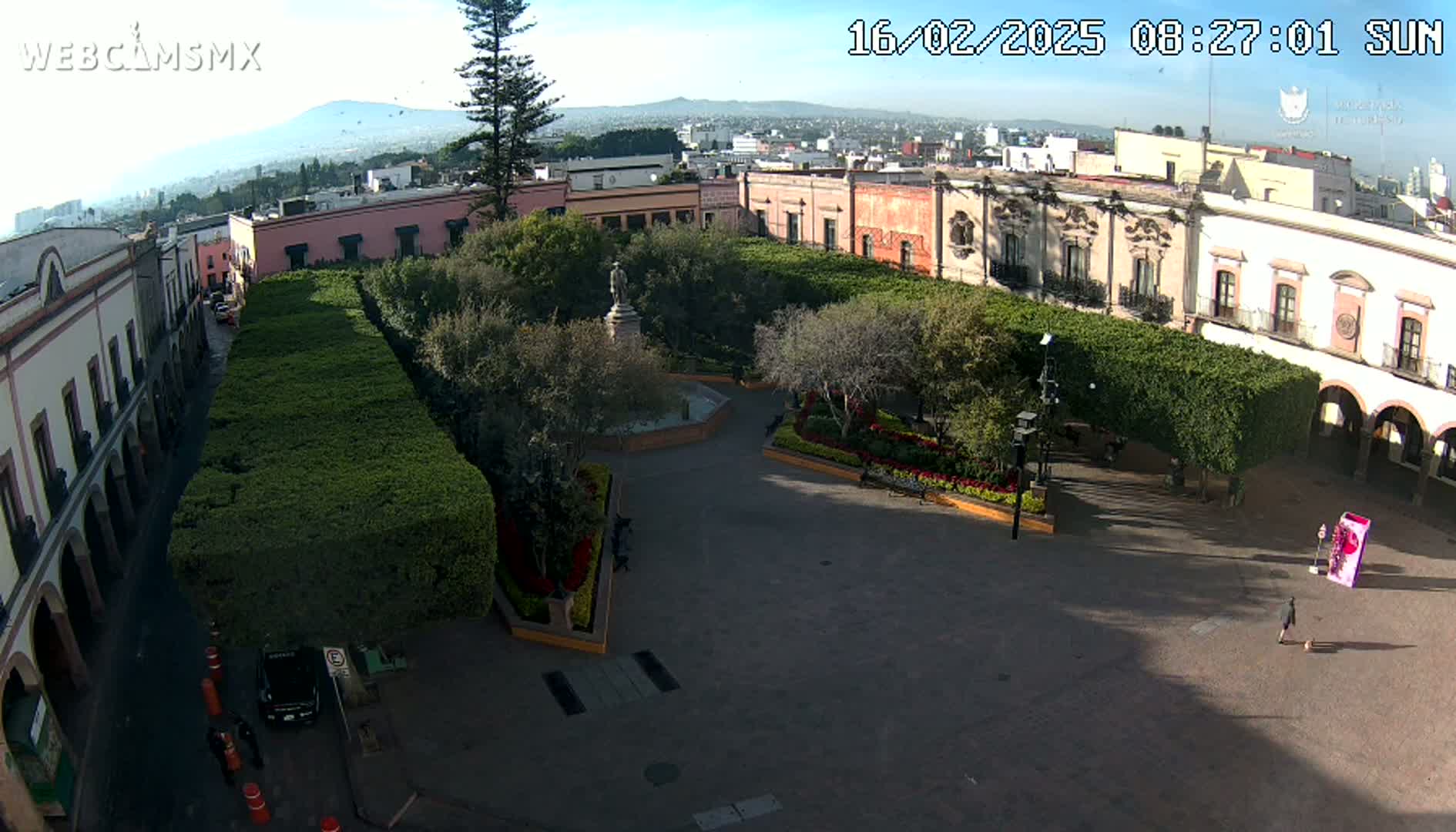 A high-angle, sunny view of a plaza surrounded by colonial-style buildings, featuring manicured hedges, a central fountain, and a few people.