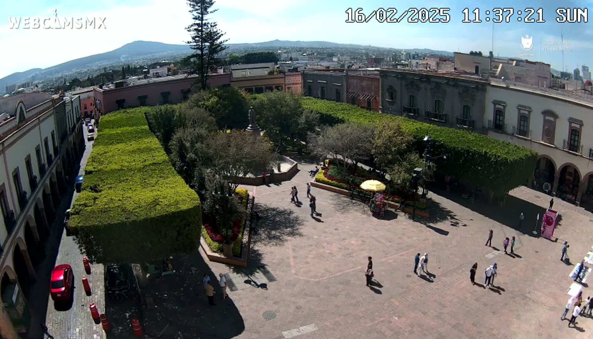 A sunny day reveals a town square with a central plaza surrounded by meticulously trimmed hedges and buildings, people walking around, and a distant mountain visible in the background.