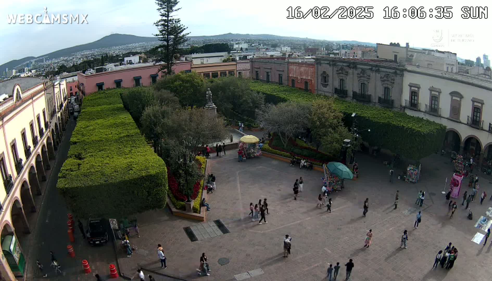 A sunny aerial view of a town square with people walking around, surrounded by colonial-style buildings and manicured hedges.