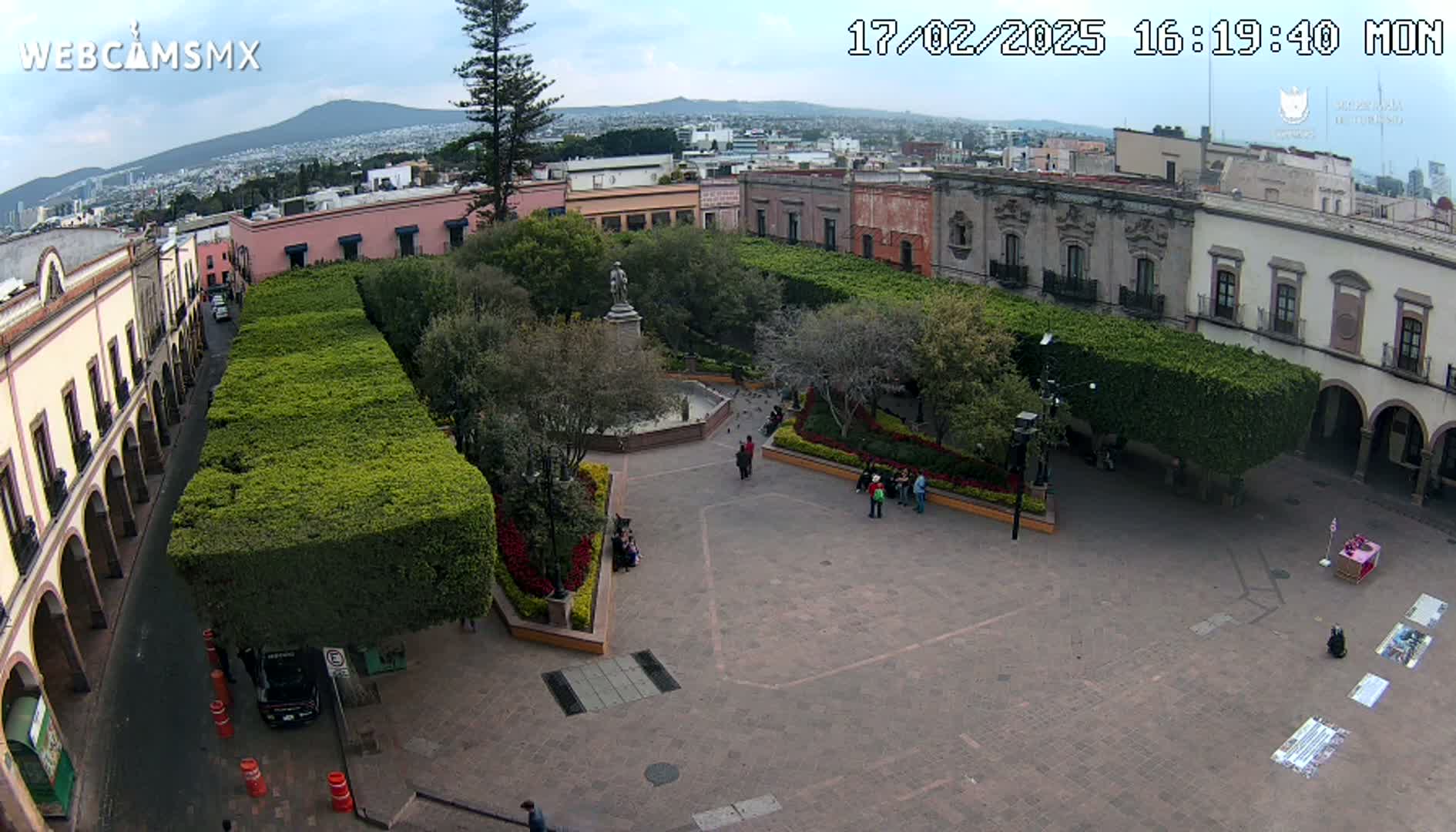 An aerial view of a town square with a central fountain and statue, surrounded by buildings and neatly trimmed hedges, under a partly cloudy sky.