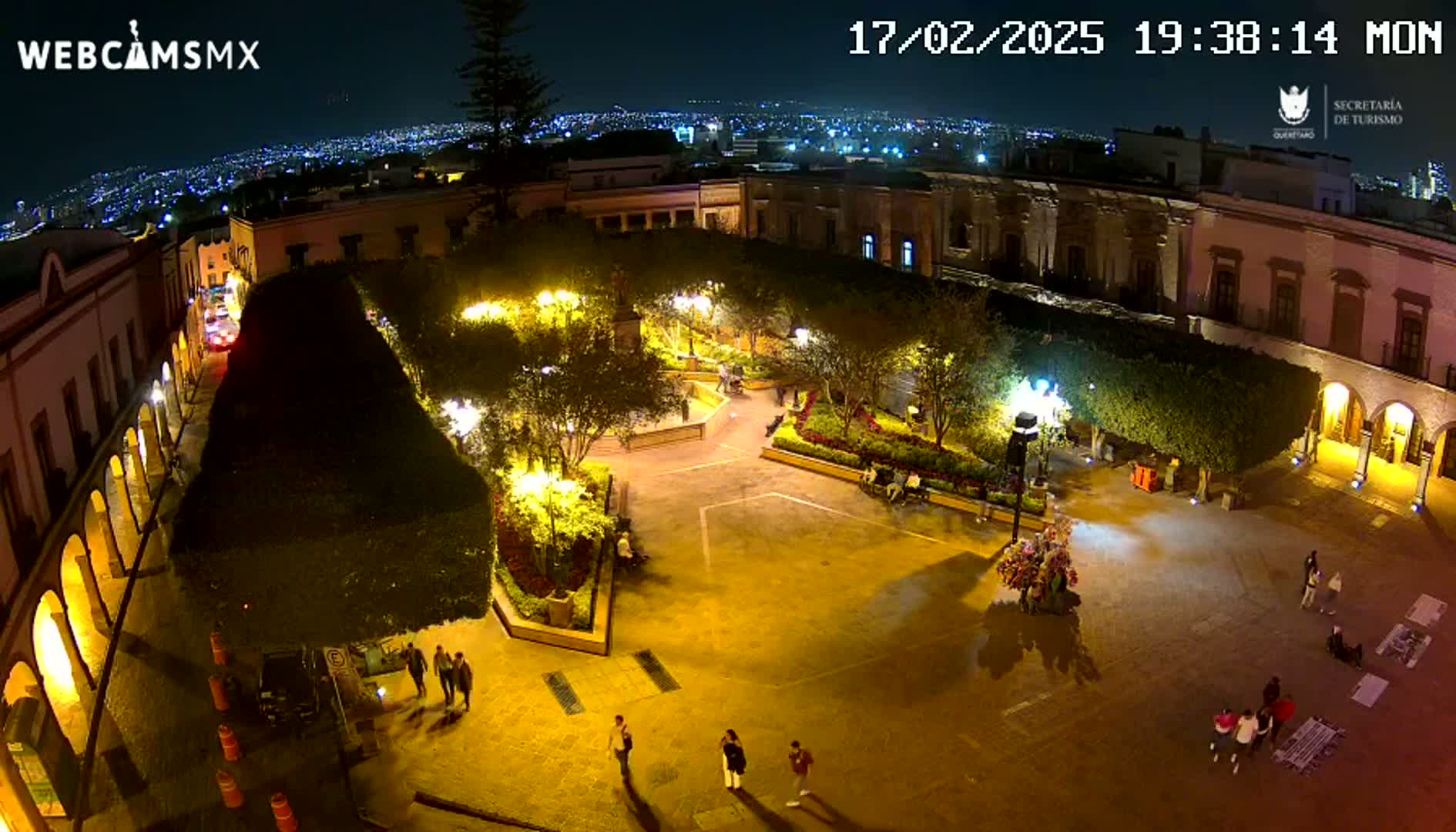An illuminated plaza at night, surrounded by buildings, with a few people walking around.