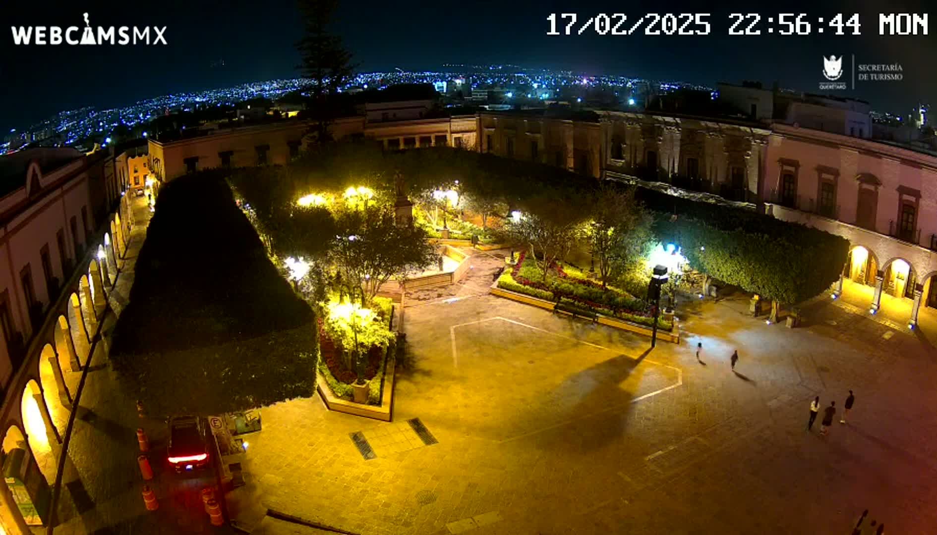 A nighttime aerial view of a brightly lit plaza surrounded by buildings, with a few people walking in the square.