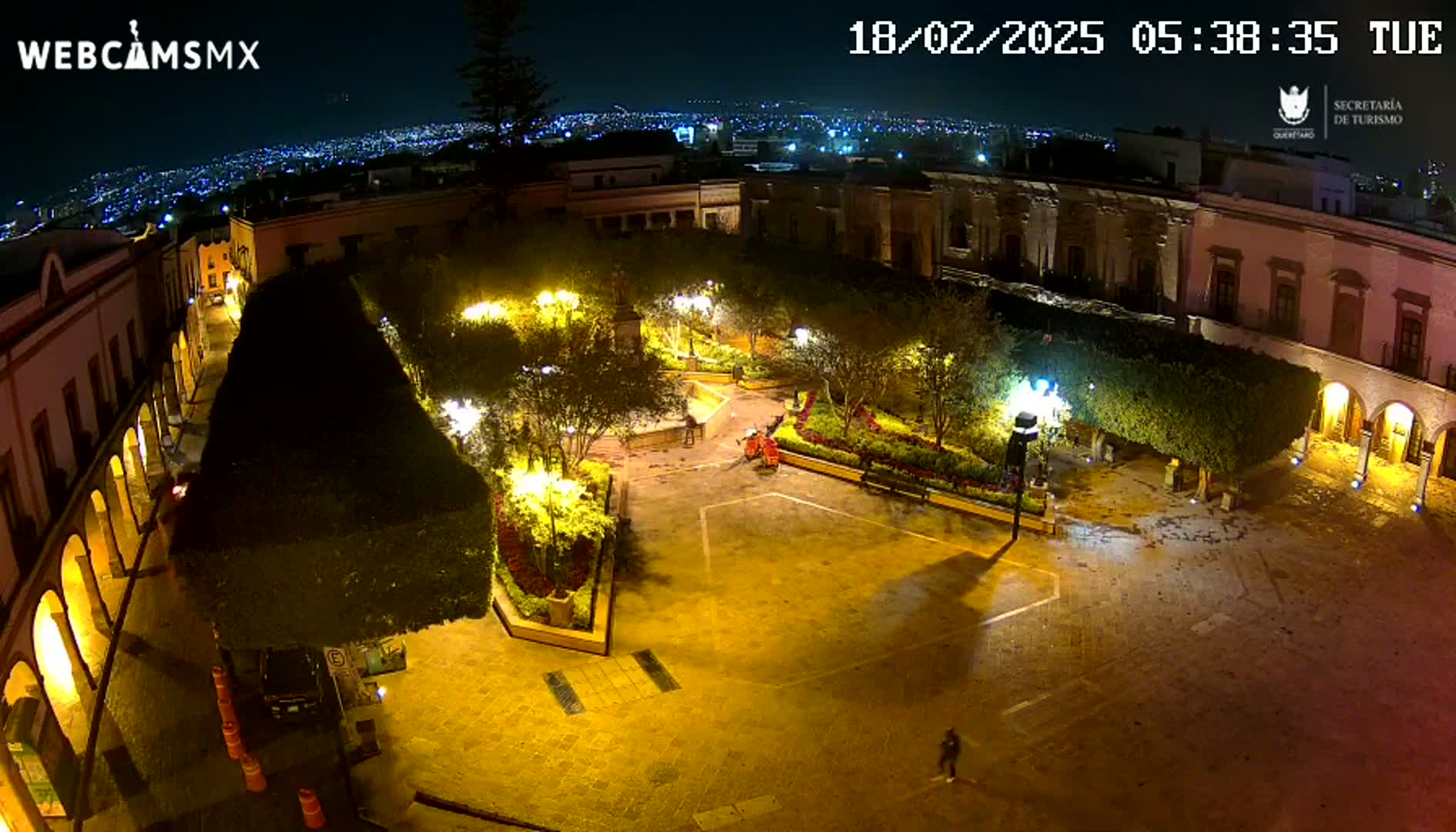 An illuminated town square at night, surrounded by buildings, with a city skyline visible in the distance under a clear sky.