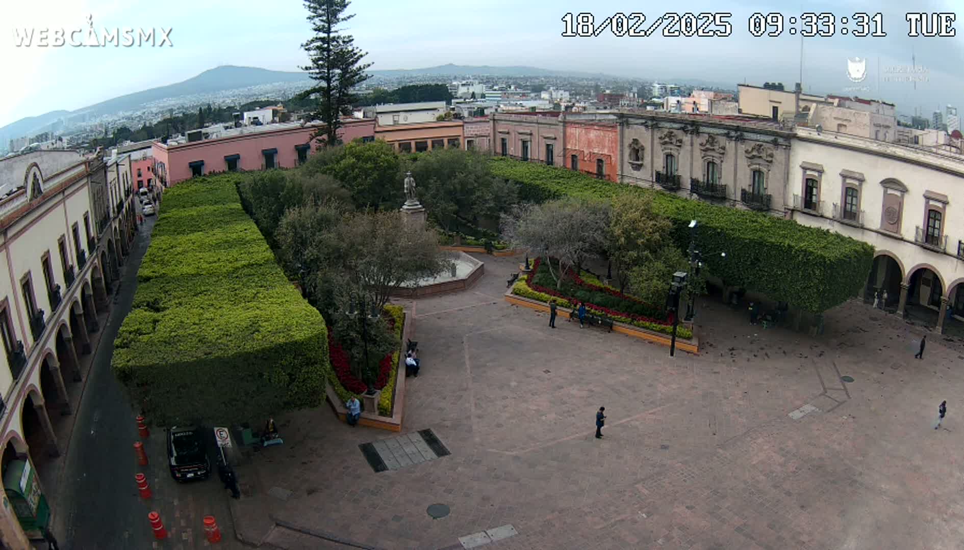 An overcast day reveals a town square with a fountain, statue, and meticulously manicured hedges, surrounded by colonial-style buildings.