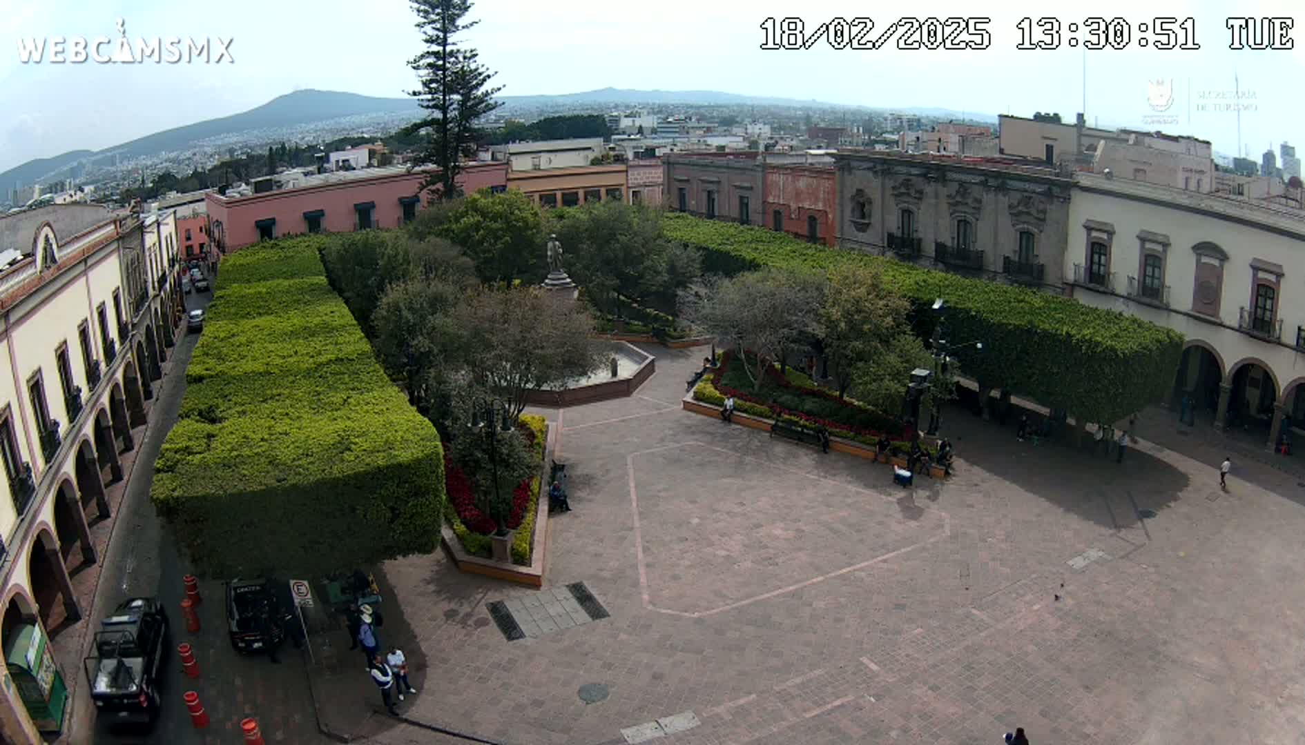 An aerial view shows a town square with a fountain and statue, surrounded by low buildings and neatly trimmed hedges under a mostly clear sky.