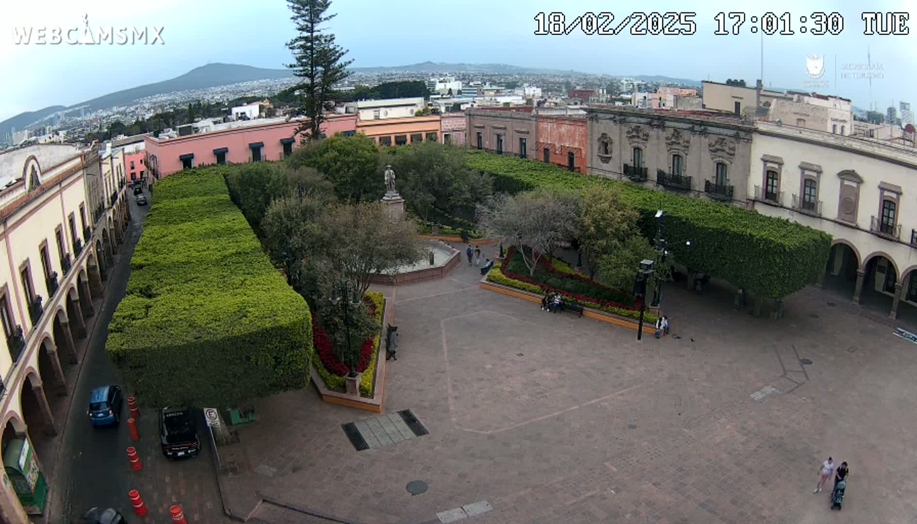 An elevated view of a town square with a central fountain and statue, surrounded by buildings and neatly trimmed hedges, under a mostly clear sky.