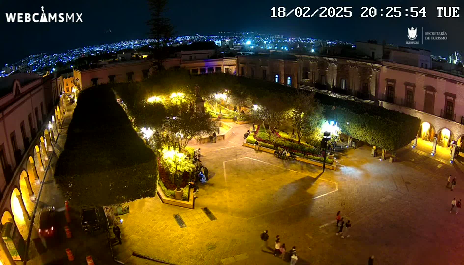 A nighttime aerial view of a town square, illuminated by streetlights, with people walking around and a city skyline visible in the distance.