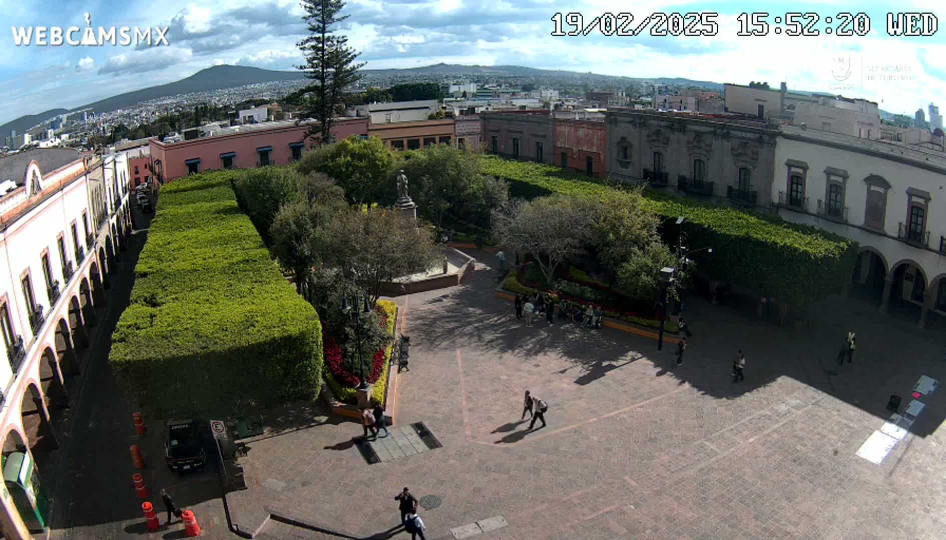 An aerial view shows a town square with low buildings, trimmed hedges, a central plaza, and a few people walking around under partly cloudy skies.