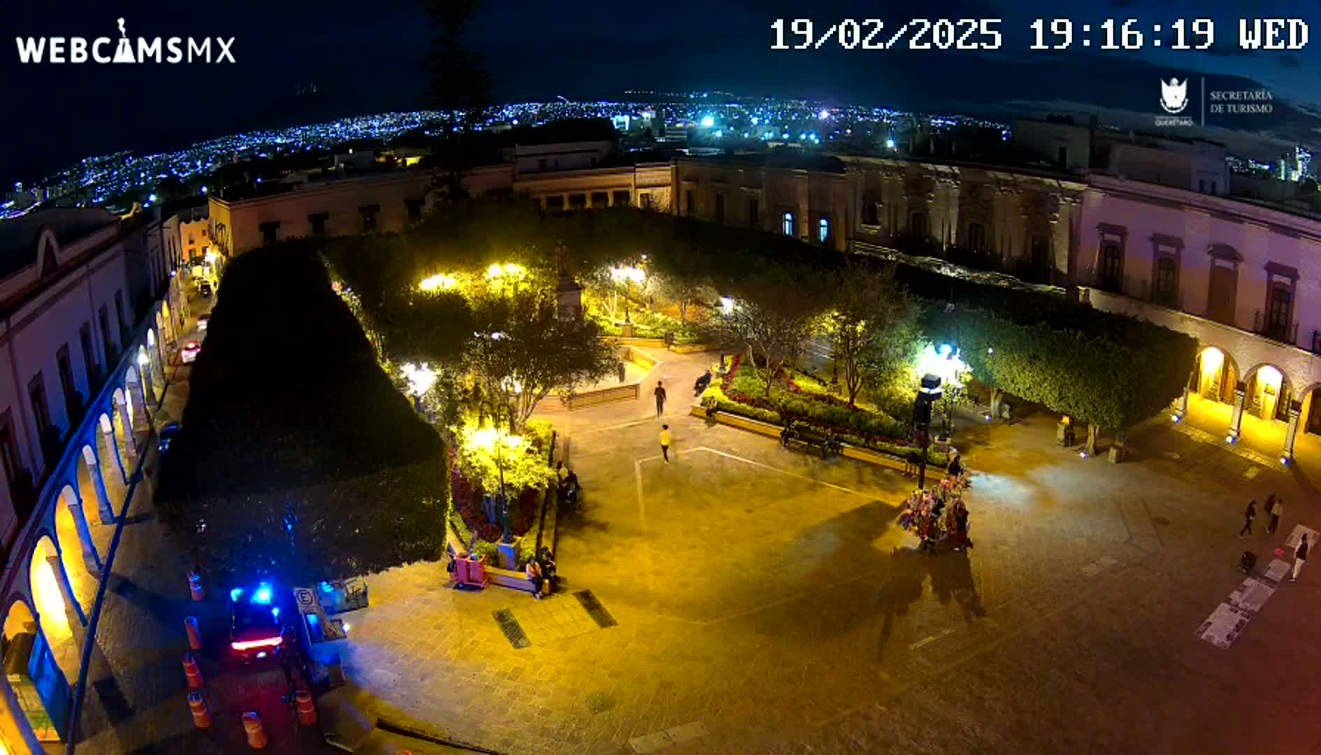 An illuminated plaza at night, surrounded by buildings, with a few people walking around and a city skyline visible in the distance.
