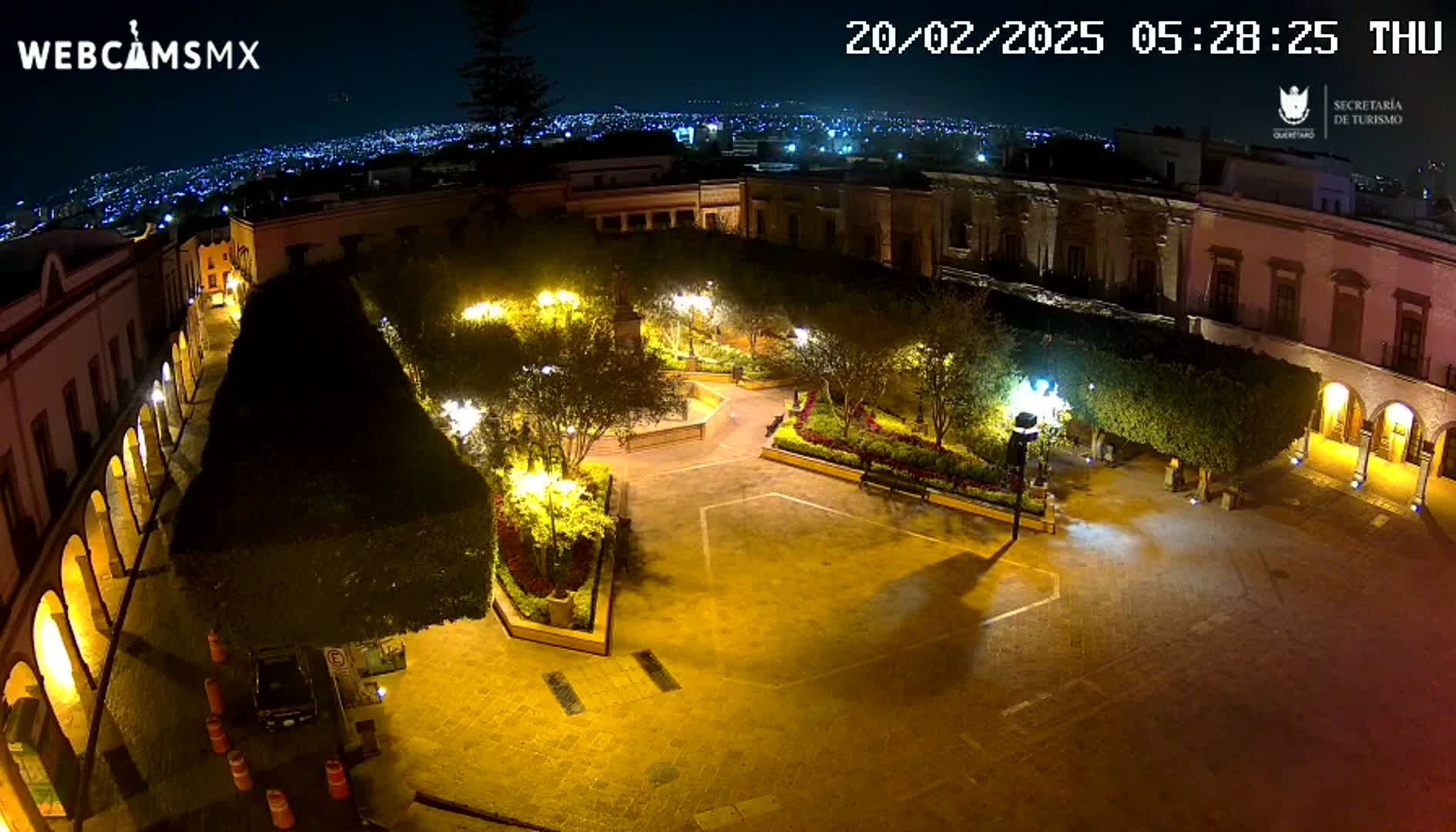 A nighttime, aerial view of a town square illuminated by lampposts, surrounded by buildings and landscaping.