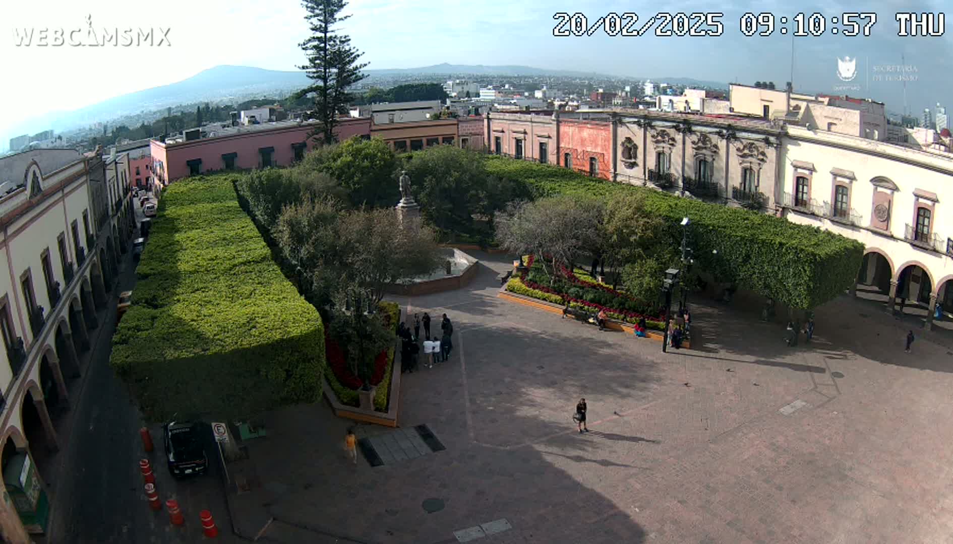 A high-angle view of a town square with a monument, surrounded by buildings and neatly trimmed hedges, under a mostly sunny sky.