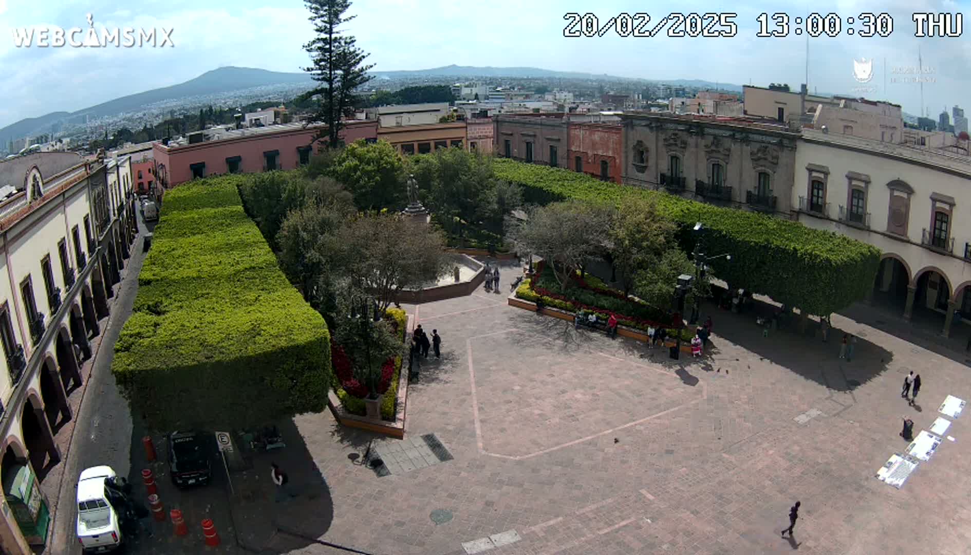 A paved plaza, surrounded by colonial-style buildings and hedges, is seen from above on a partly sunny day, with a distant mountain visible in the background.