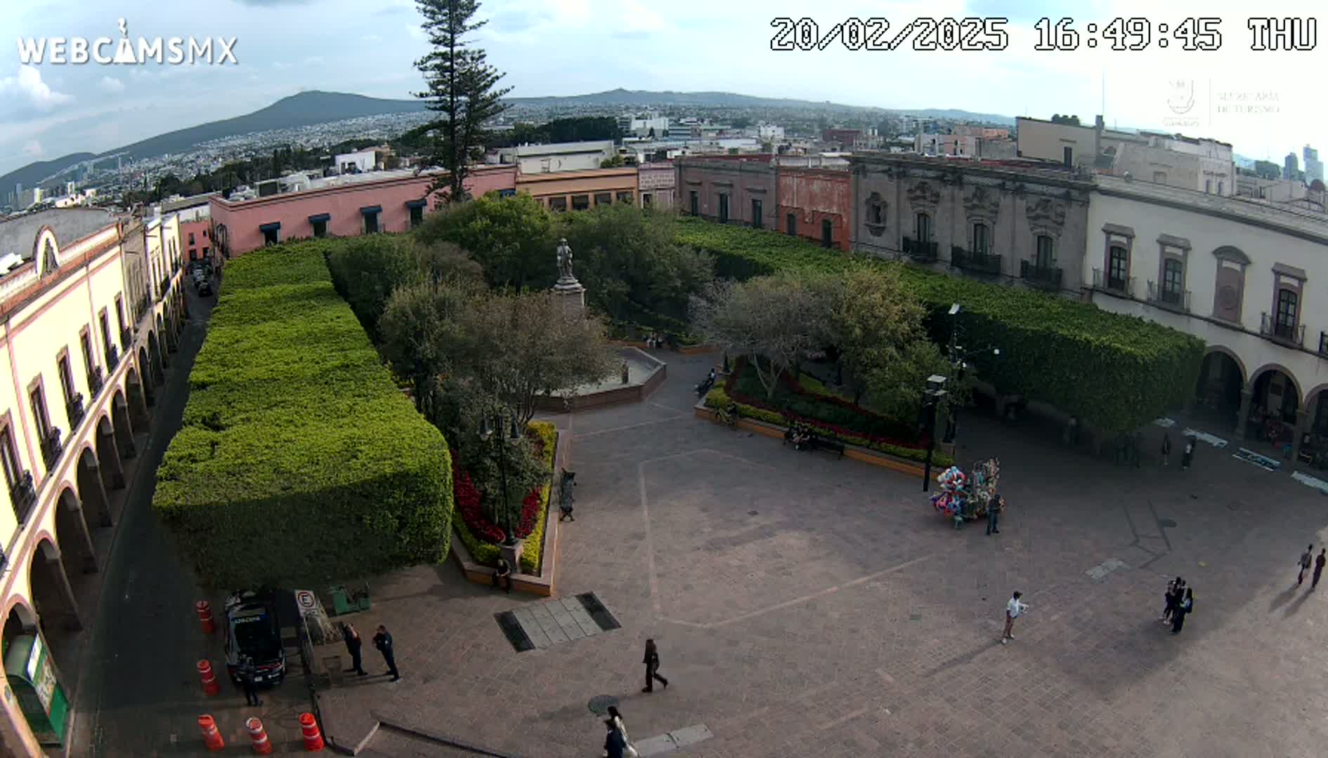 An aerial view of a town square, under partly cloudy skies, shows a plaza surrounded by buildings and trimmed hedges, with a few people walking around.