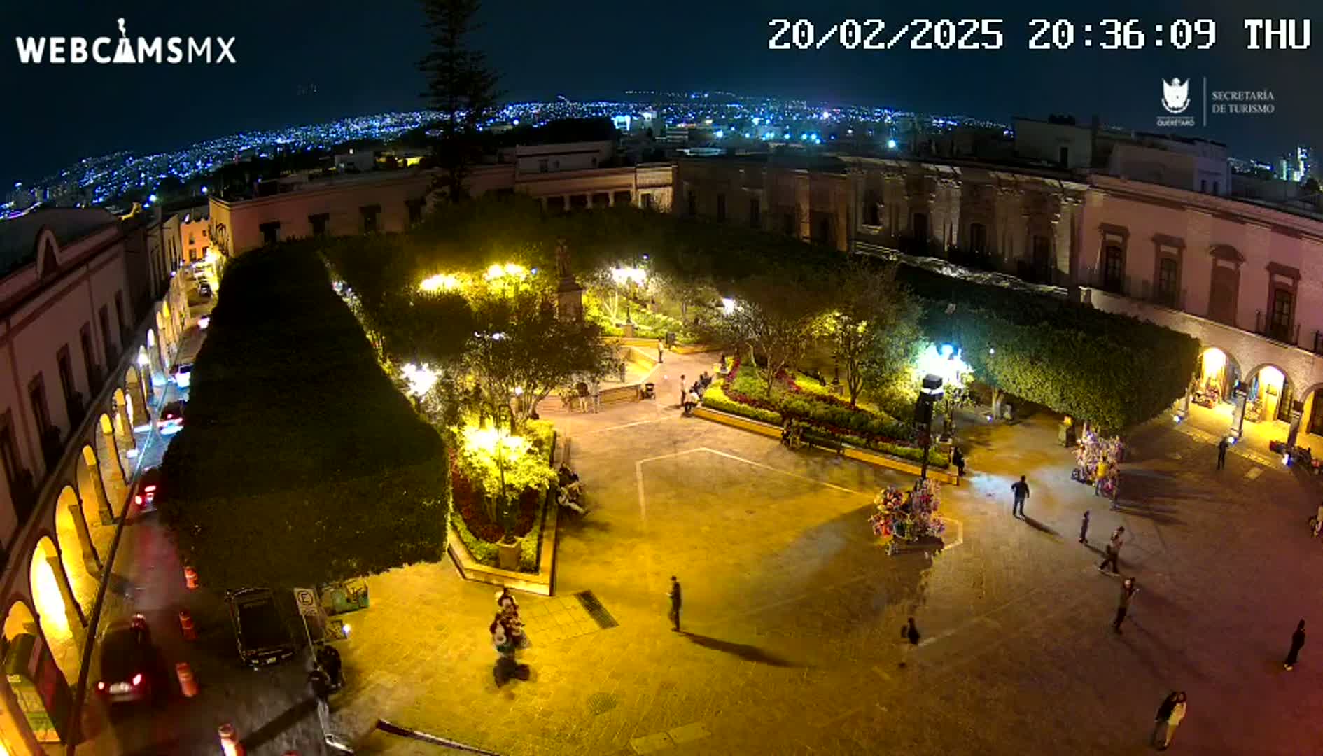 A nighttime, illuminated plaza in a city, surrounded by buildings, with people walking around and a city skyline visible in the background.