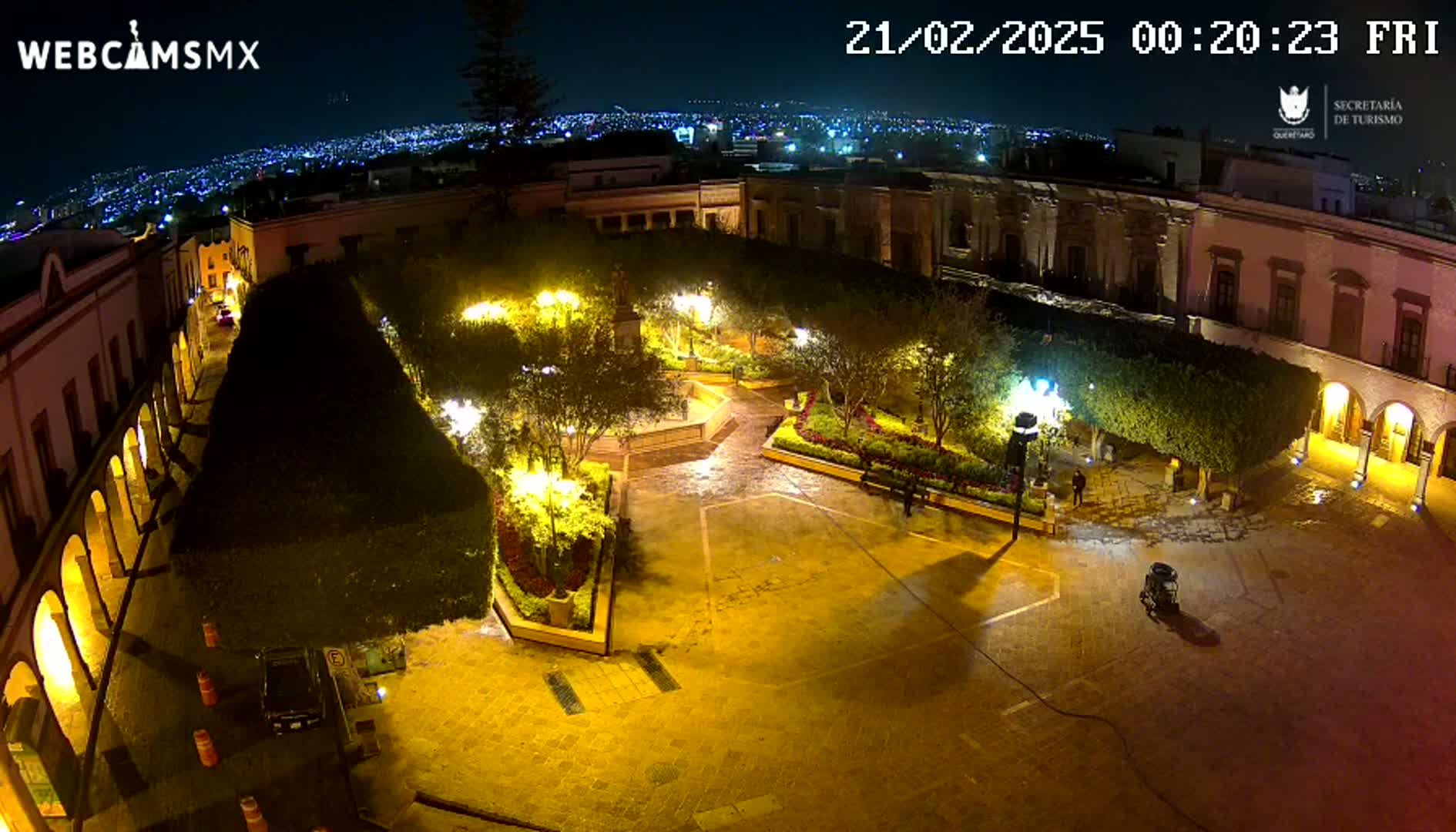 A nighttime, illuminated plaza with a city view in the background, slightly wet from rain.