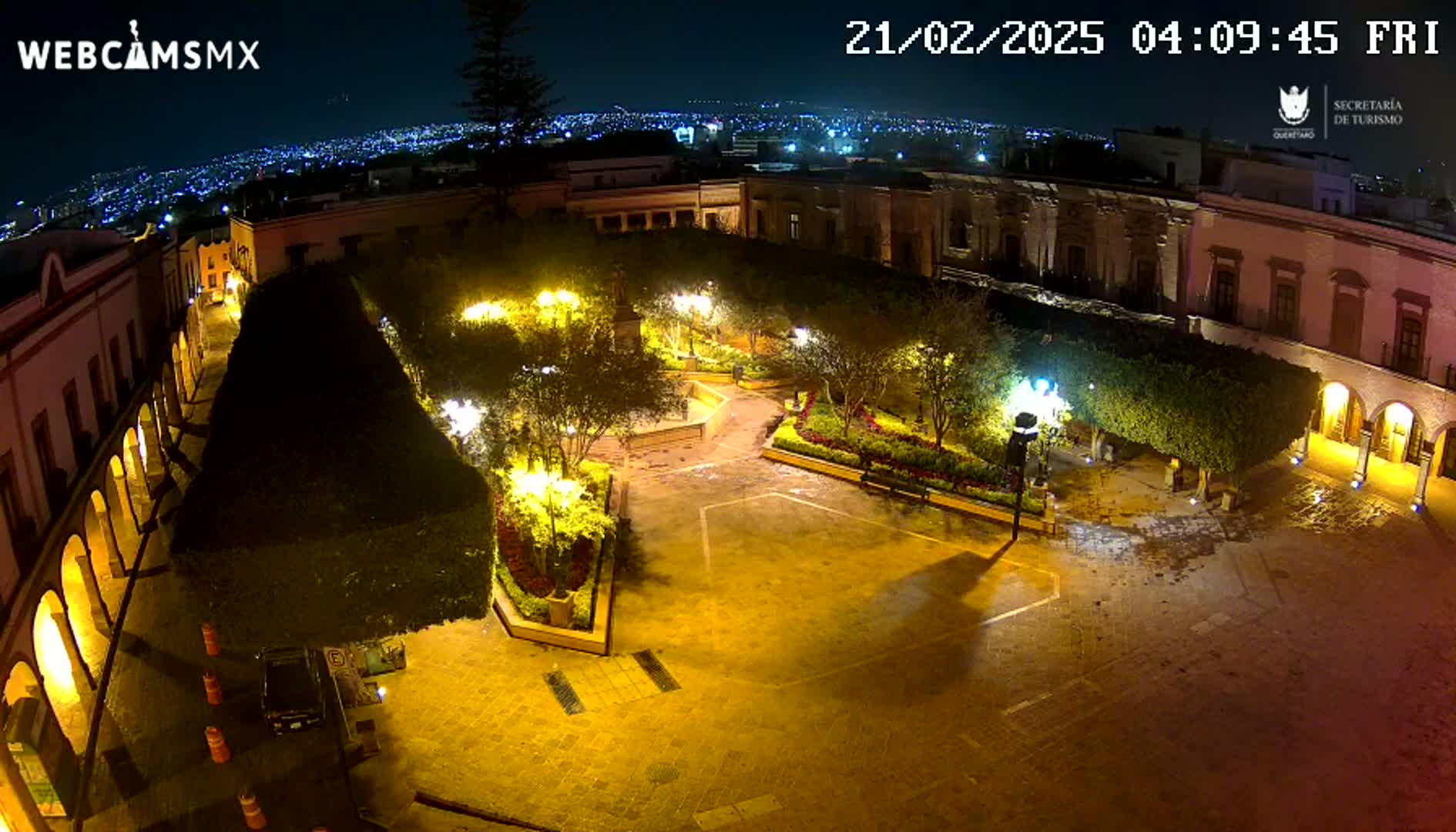 A nighttime, overhead view of a town square, illuminated by streetlights, with buildings surrounding a paved plaza containing landscaped areas and a few parked vehicles.