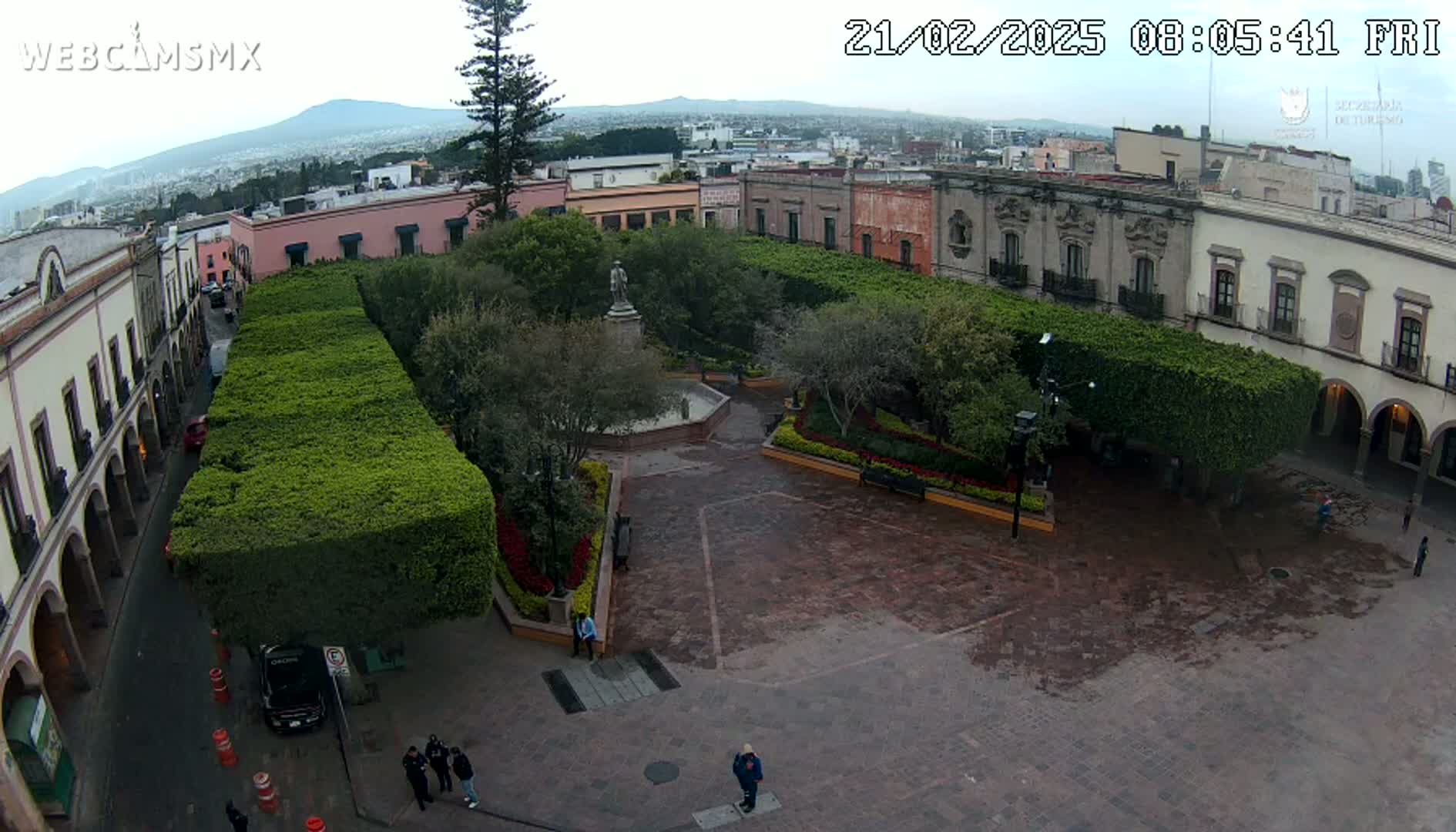 An overcast day reveals a town square with a central fountain, surrounded by neatly trimmed hedges, colonial-style buildings, and a few pedestrians.