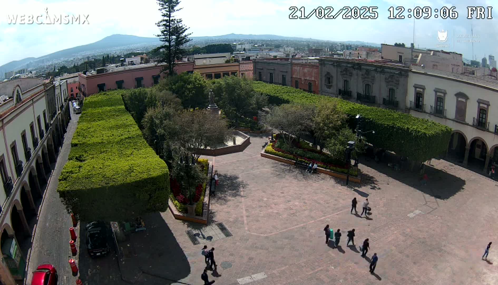 An aerial view of a town square with a central plaza, surrounded by buildings and neatly trimmed hedges, on a sunny day with a mountain visible in the distance.