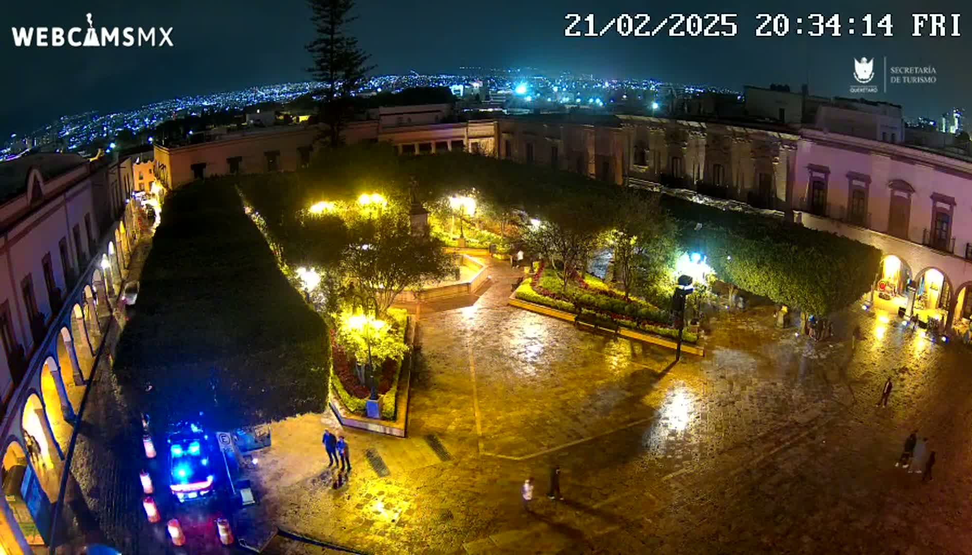 A rain-slicked, illuminated plaza at night, surrounded by colonial-style buildings, with a few people visible.