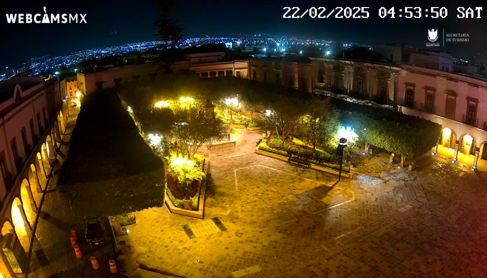 A nighttime aerial view of a plaza, illuminated by streetlights, surrounded by buildings, with a city skyline visible in the distance under a clear sky.