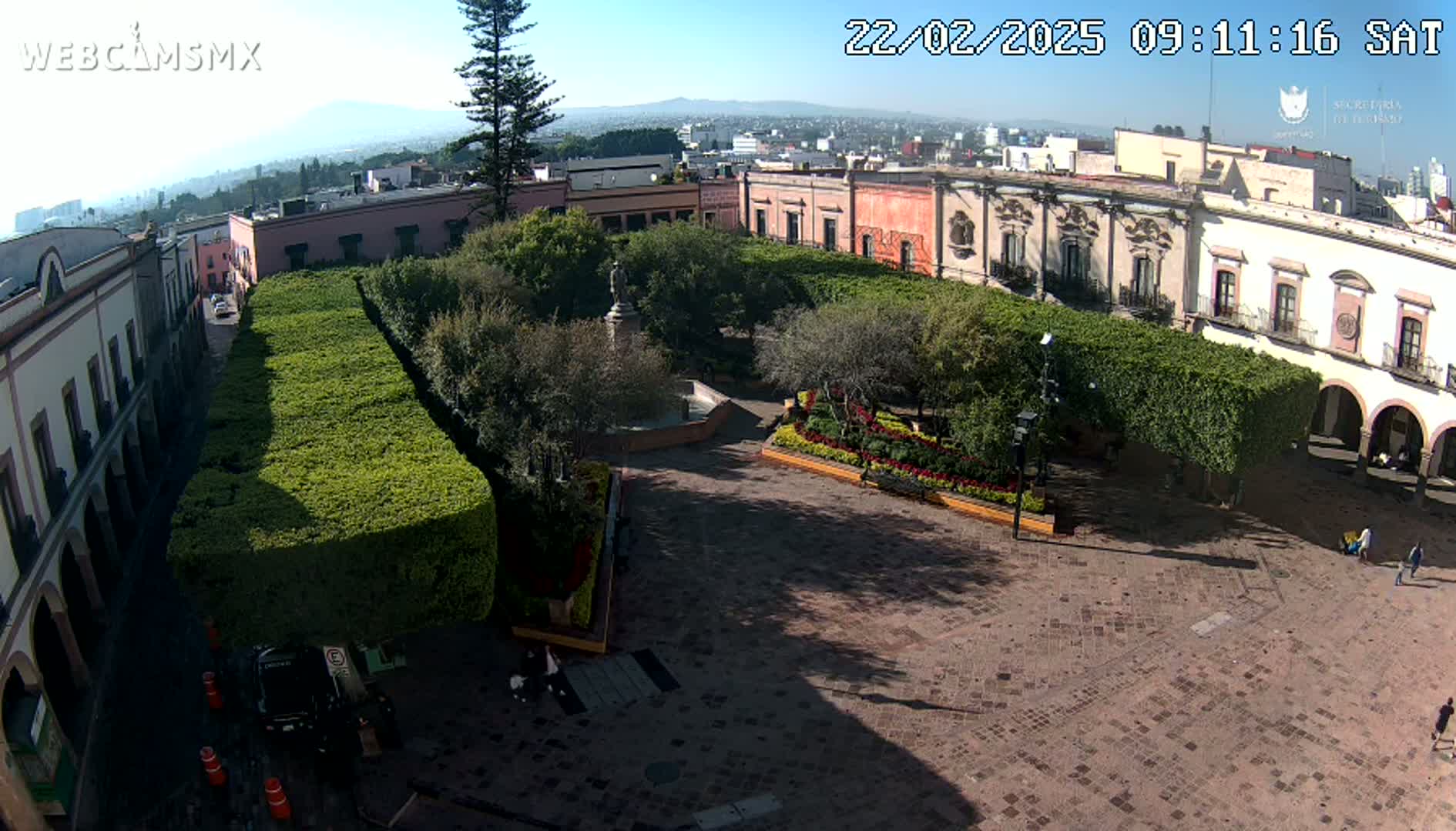 A sunny day reveals a plaza with meticulously manicured hedges, a central fountain, and colonial-era buildings surrounding it.