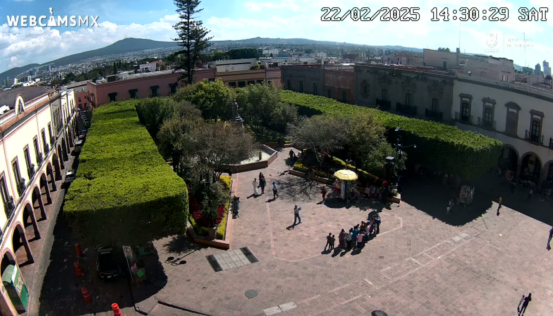 An elevated view of a town square with a central plaza surrounded by buildings and lush greenery under a sunny sky.