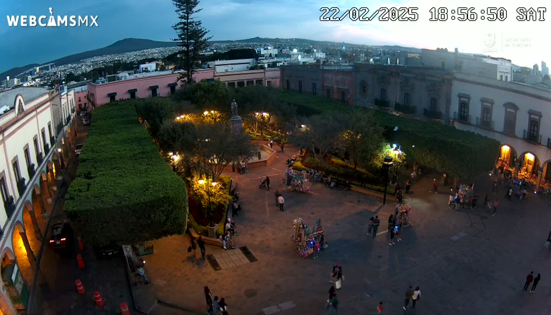 An aerial evening view of a town square, illuminated with lights, surrounded by buildings and lush green hedges, under a twilight sky.