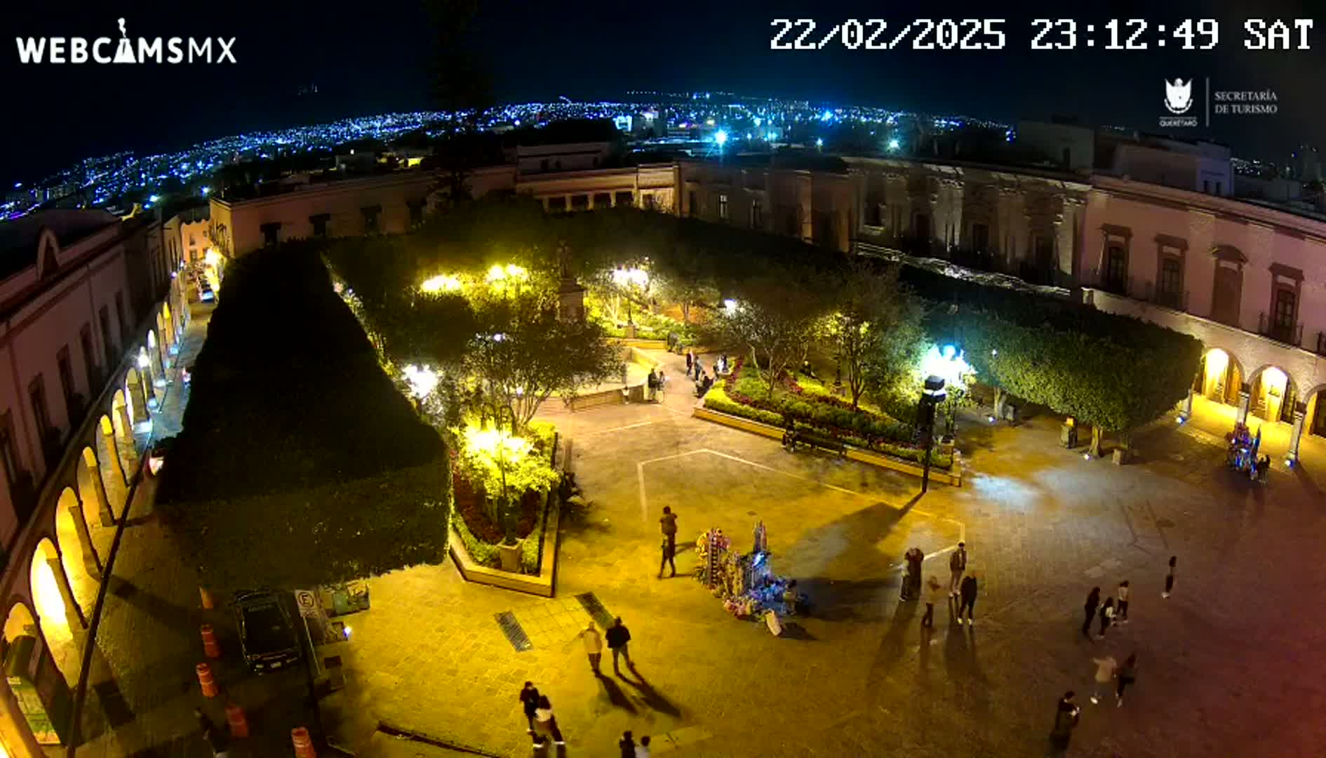 A nighttime aerial view of a town square, illuminated by streetlights, with people walking around and a city skyline visible in the background under a clear sky.