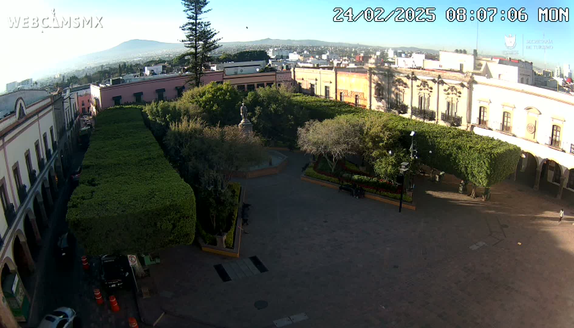 A sunny day reveals a town square with a statue in the center, surrounded by neatly trimmed hedges, and colonial-style buildings.