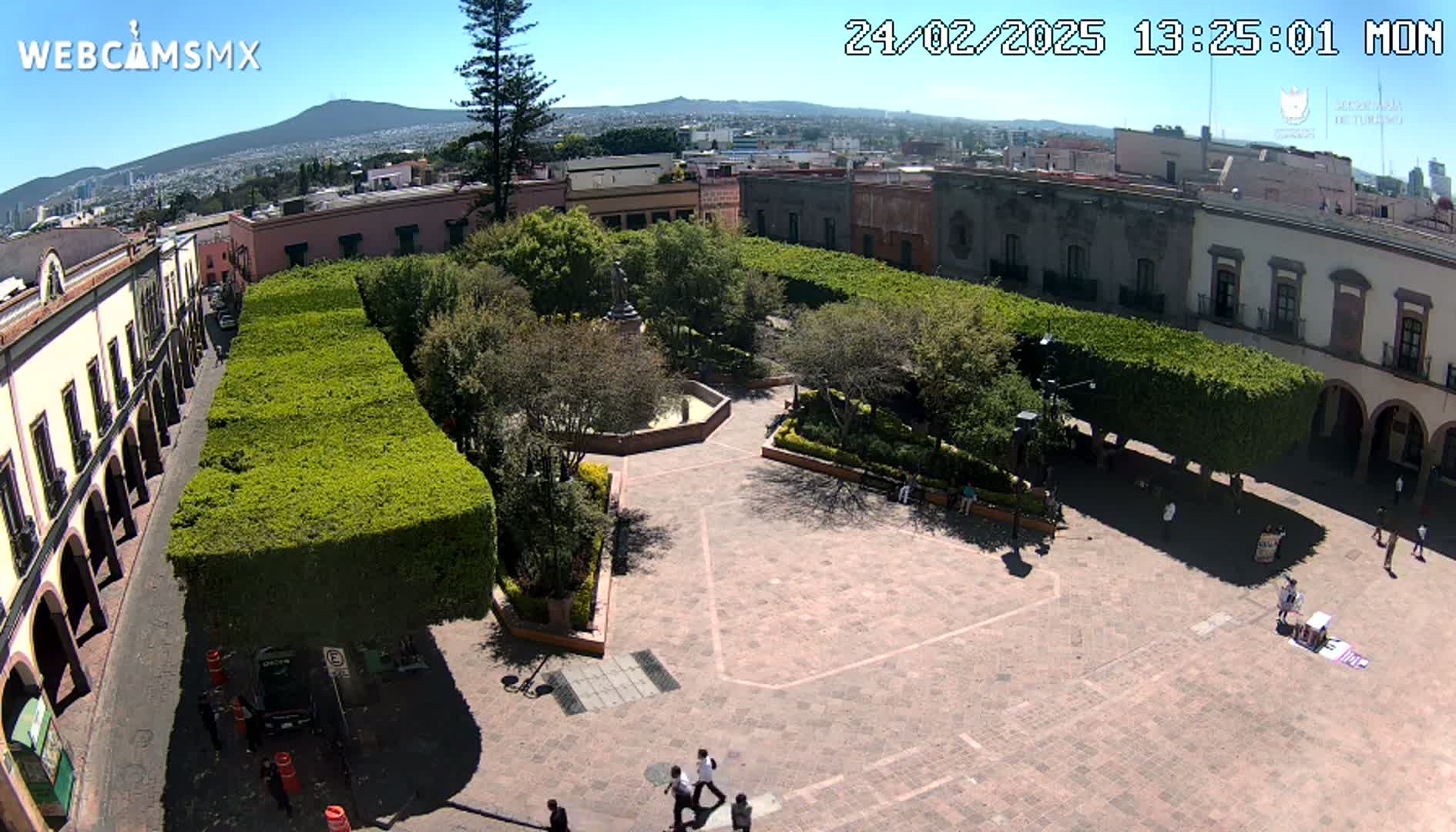 A sunny day reveals a town square with a central plaza surrounded by trimmed hedges, buildings, and a few people walking around.