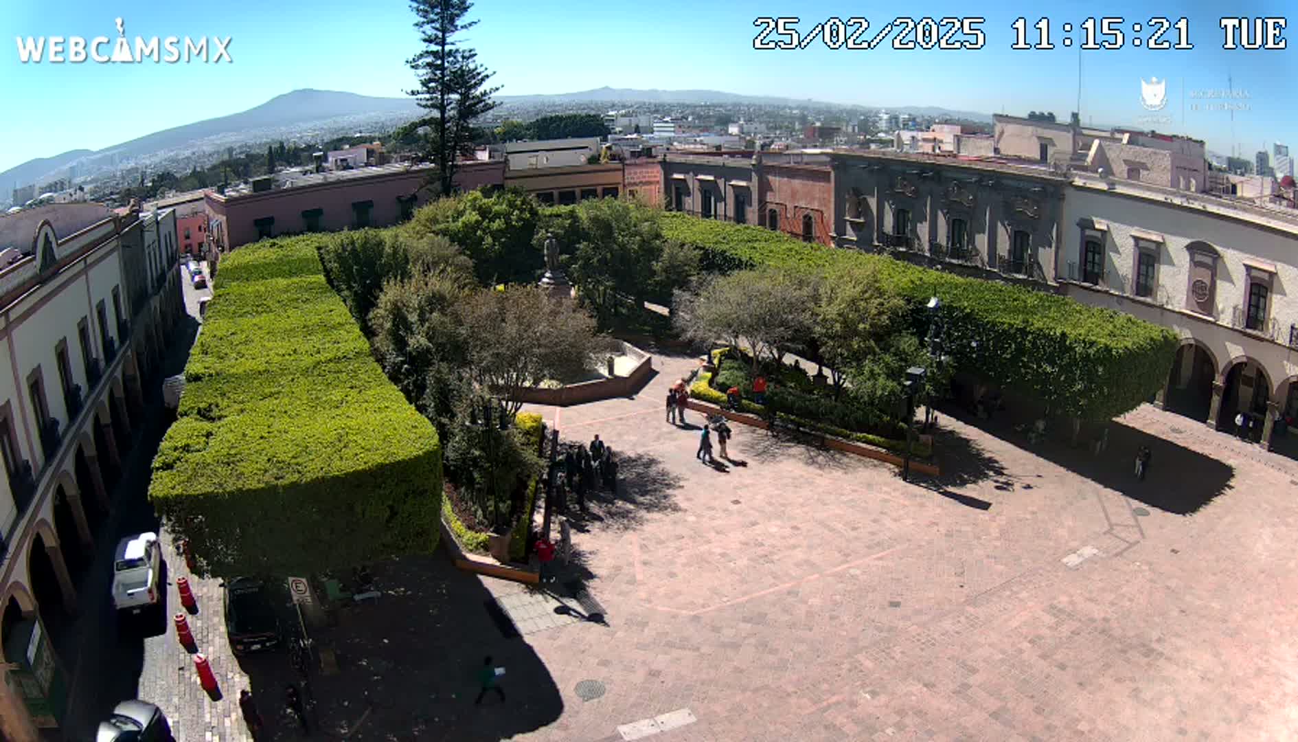 A sunny day reveals a town square with a central park featuring neatly trimmed hedges, a fountain, and several people walking around, all surrounded by colonial-style buildings, and a distant mountain range is visible in the background.