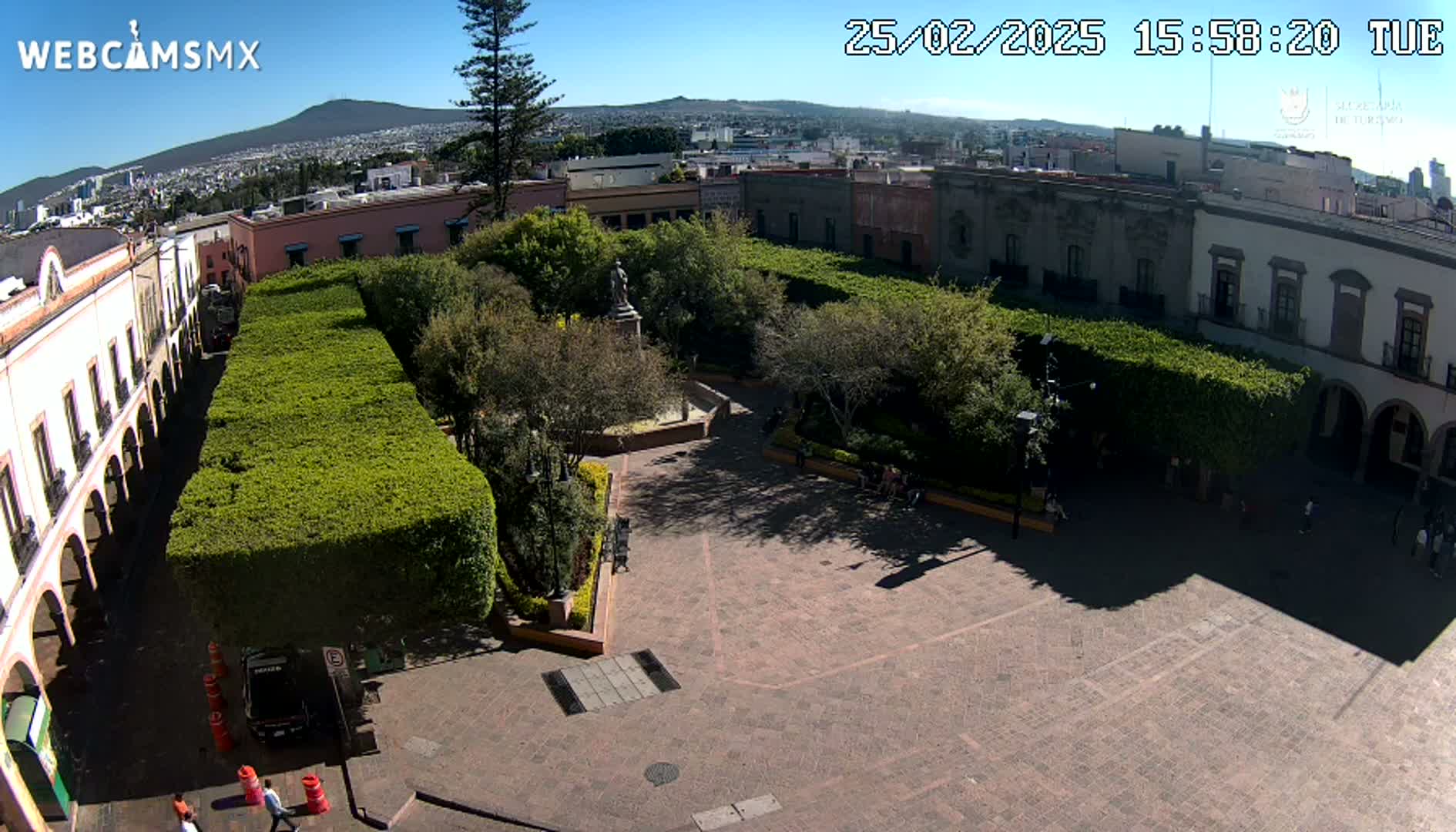 An aerial view of a plaza with a central garden, surrounded by colonial-style buildings under a clear, sunny sky.