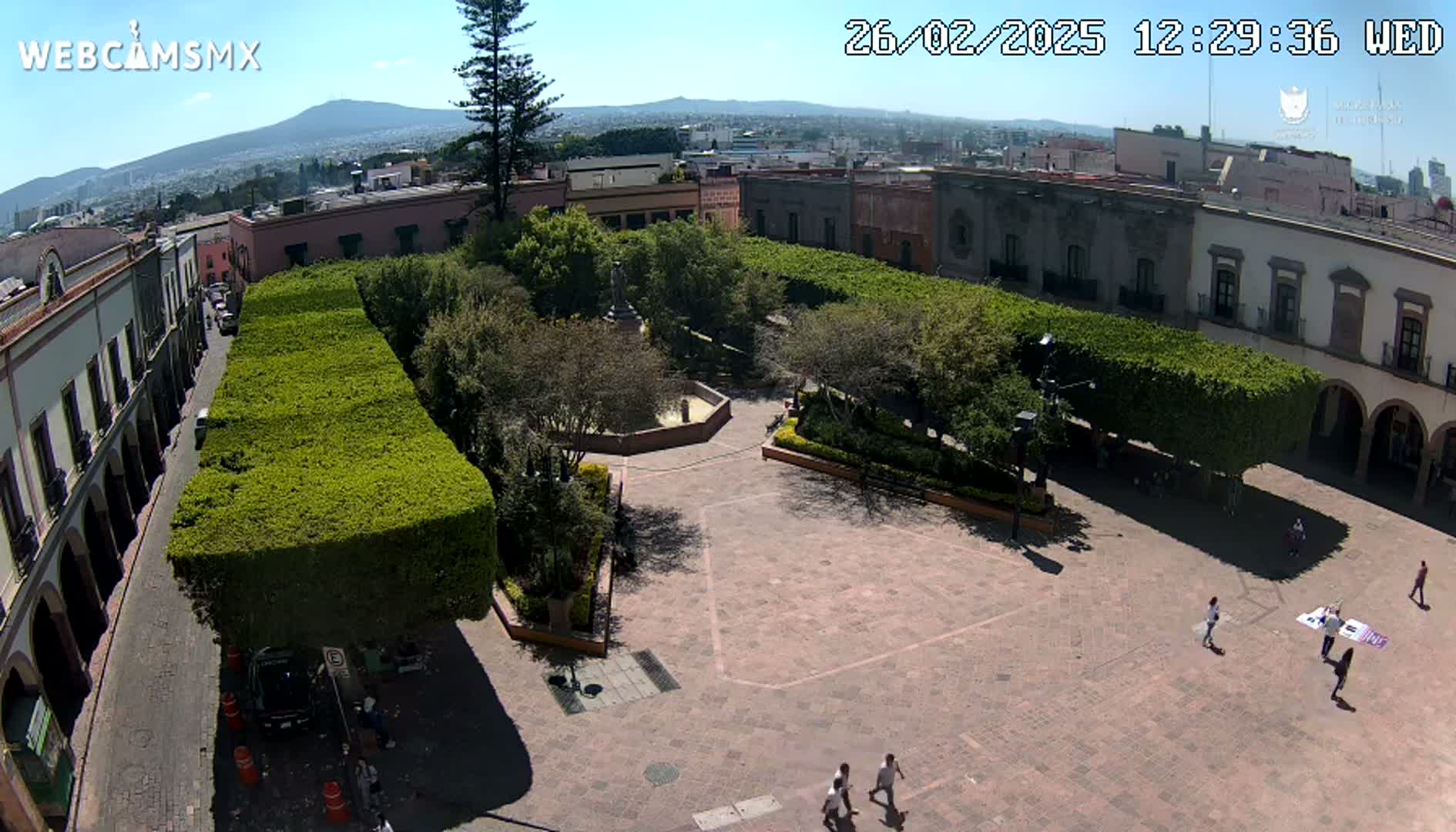 A sunny aerial view of a town square with a central fountain, surrounded by buildings and meticulously trimmed hedges.