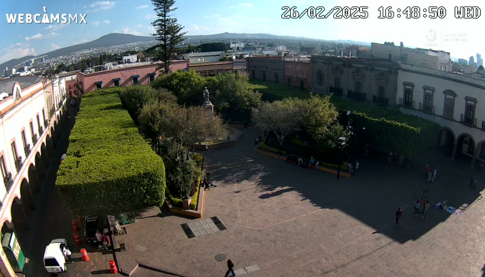 A sunny day reveals a plaza with a central statue, surrounded by trimmed hedges, colonial-style buildings, and a few people scattered about.