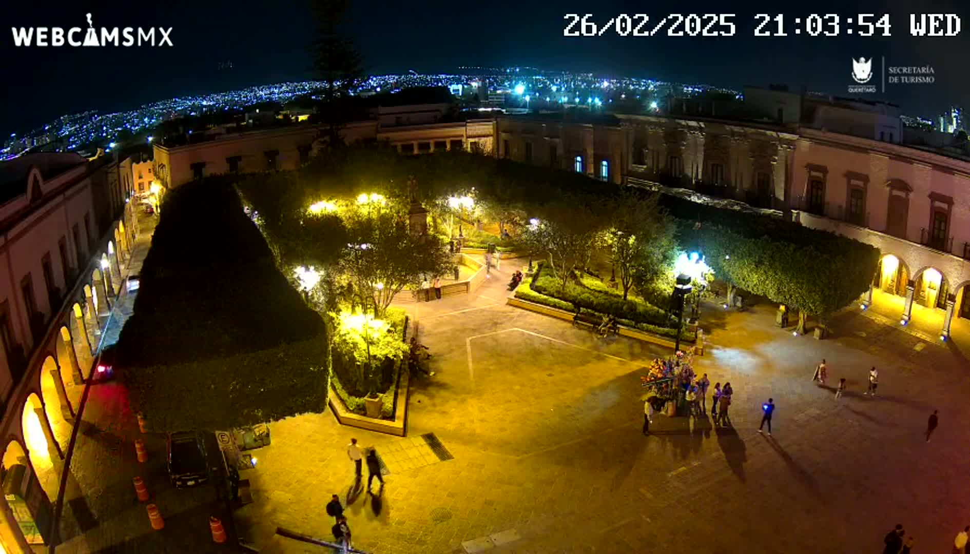 A nighttime aerial view of a brightly lit plaza surrounded by buildings, with a few people walking around.