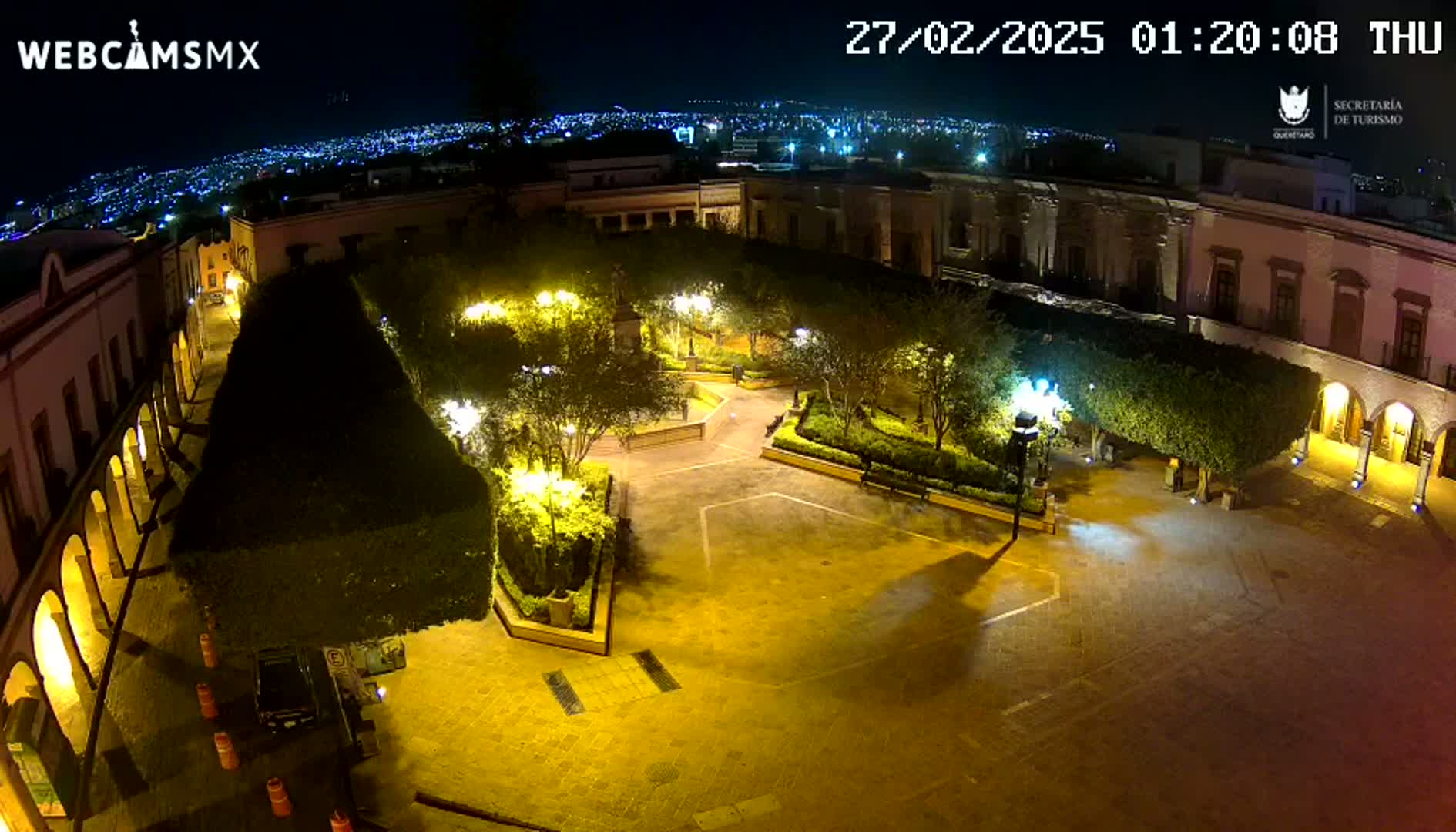 A nighttime aerial view of a town square, illuminated by streetlights, surrounded by buildings and landscaped gardens.