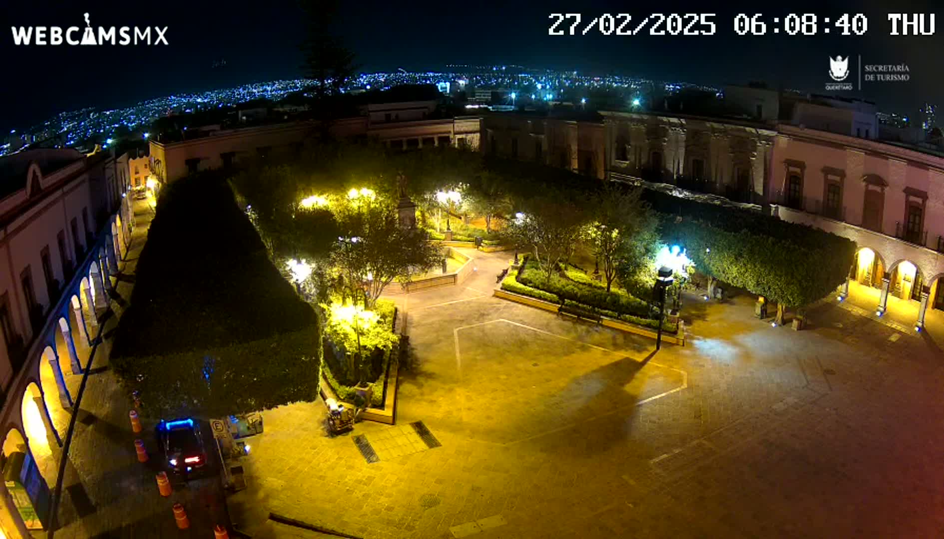 A brightly lit plaza at night, surrounded by colonial-style buildings, with a city skyline visible in the distance under a clear sky.