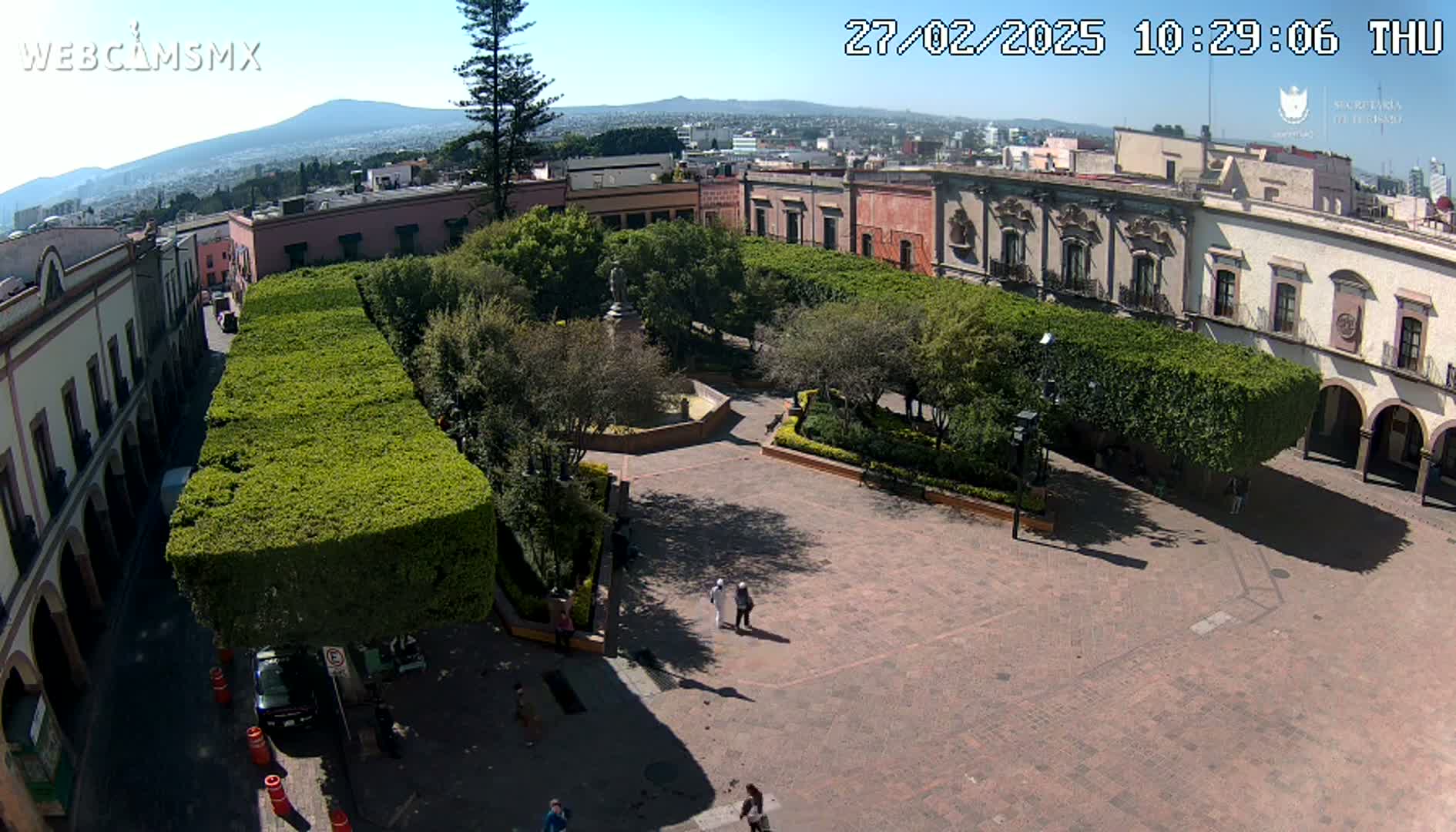 A sunny day reveals a plaza surrounded by colonial-style buildings, meticulously landscaped with hedges, and a central fountain.