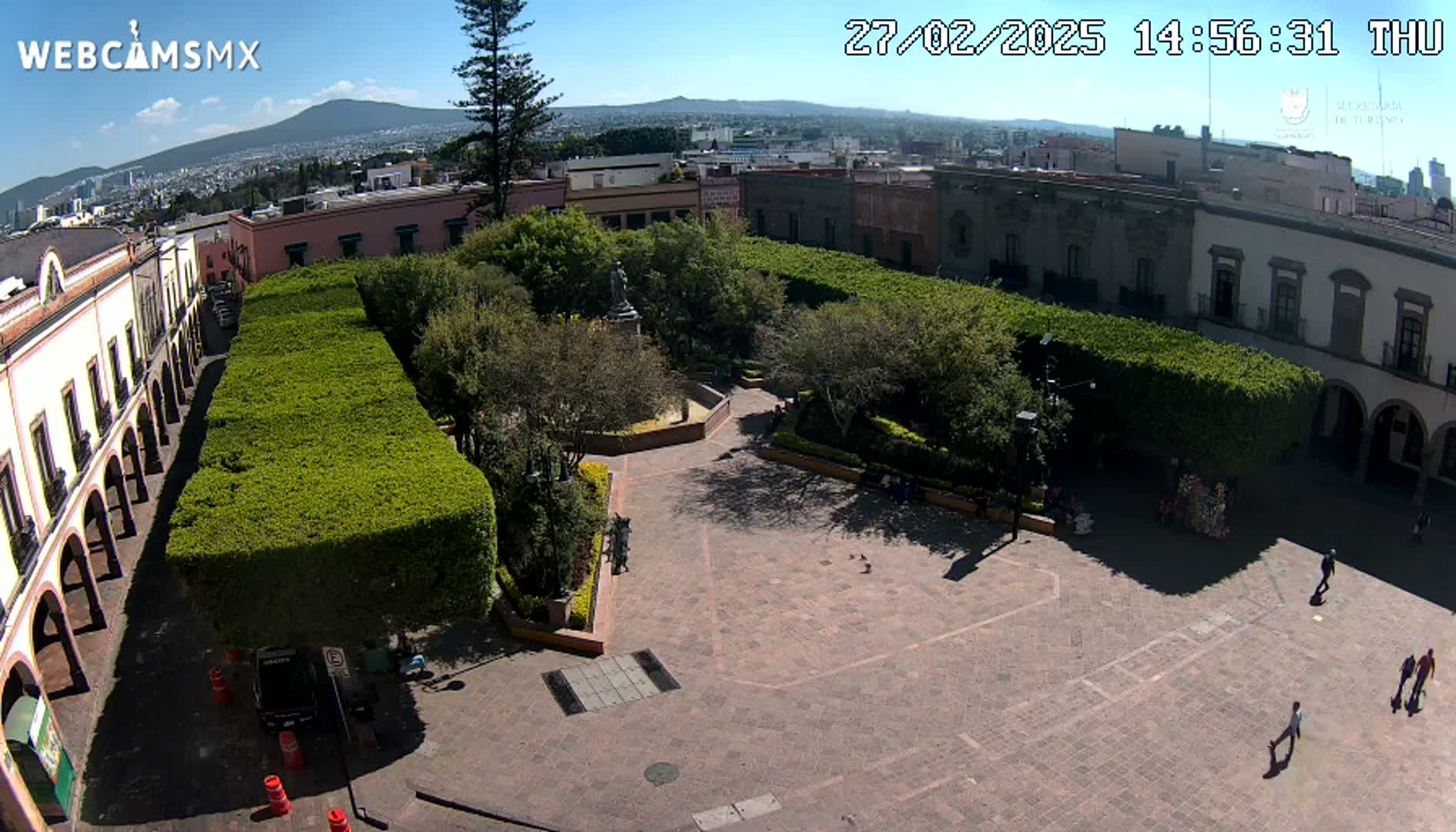 An elevated view of a town square with neatly trimmed hedges, colonial-style buildings, and a few people walking on a sunny day.